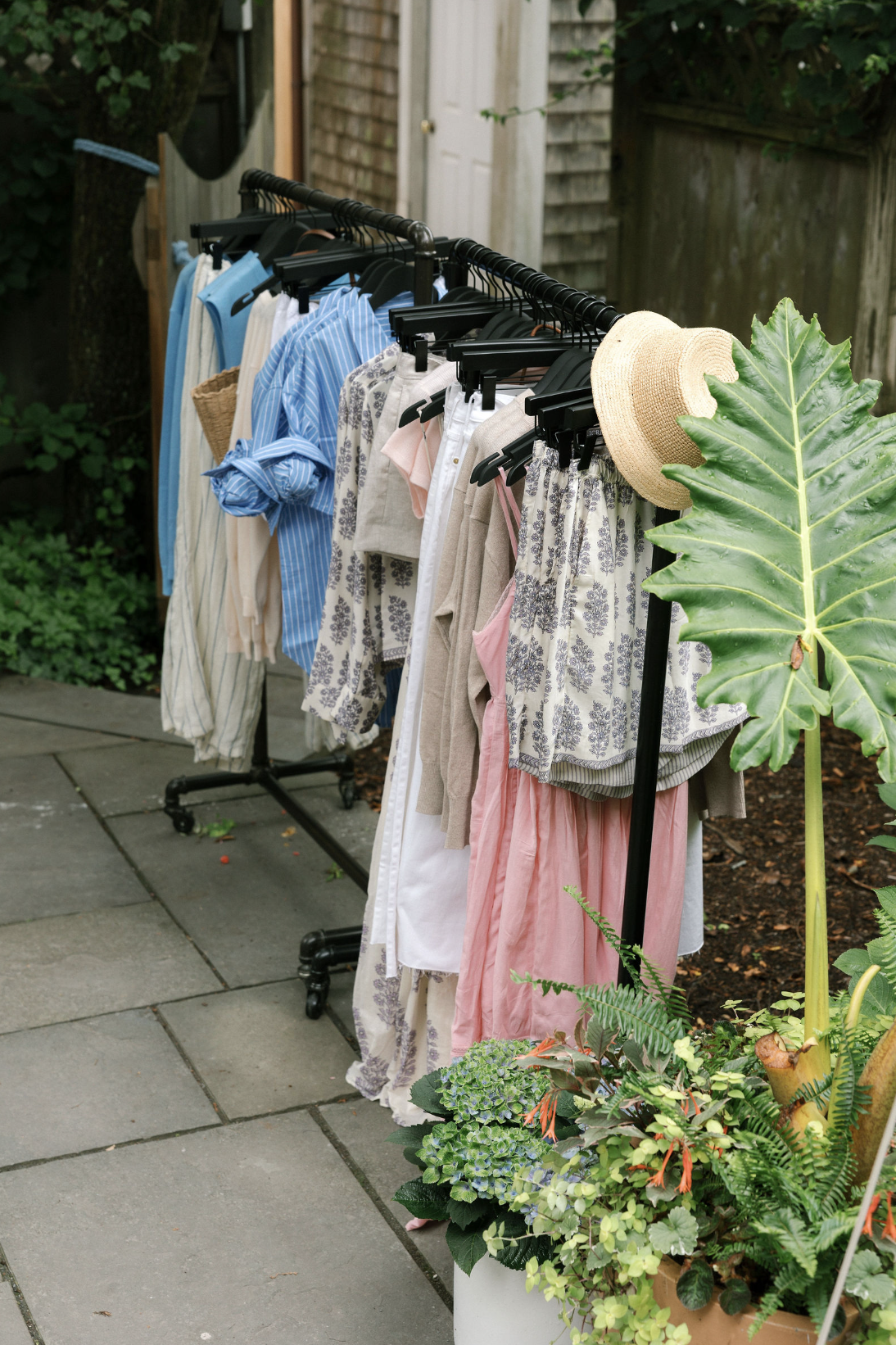 Outdoor clothing rack with dresses, shirts, and skirts, a straw hat on top, and a large green leafy plant nearby.