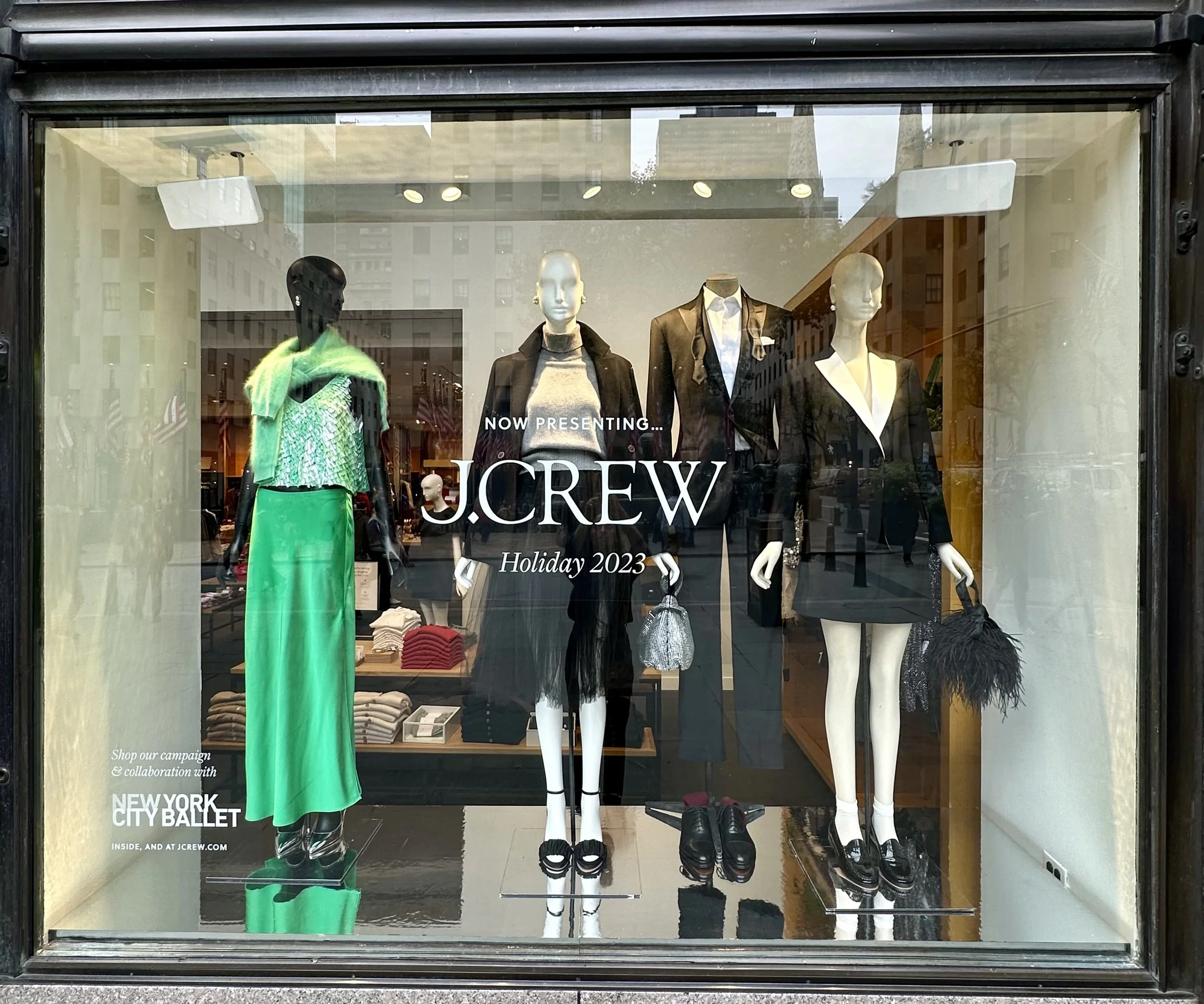 Mannequins dressed in fashionable clothing displayed in J.Crew store window with signage announcing holiday 2023 collection.