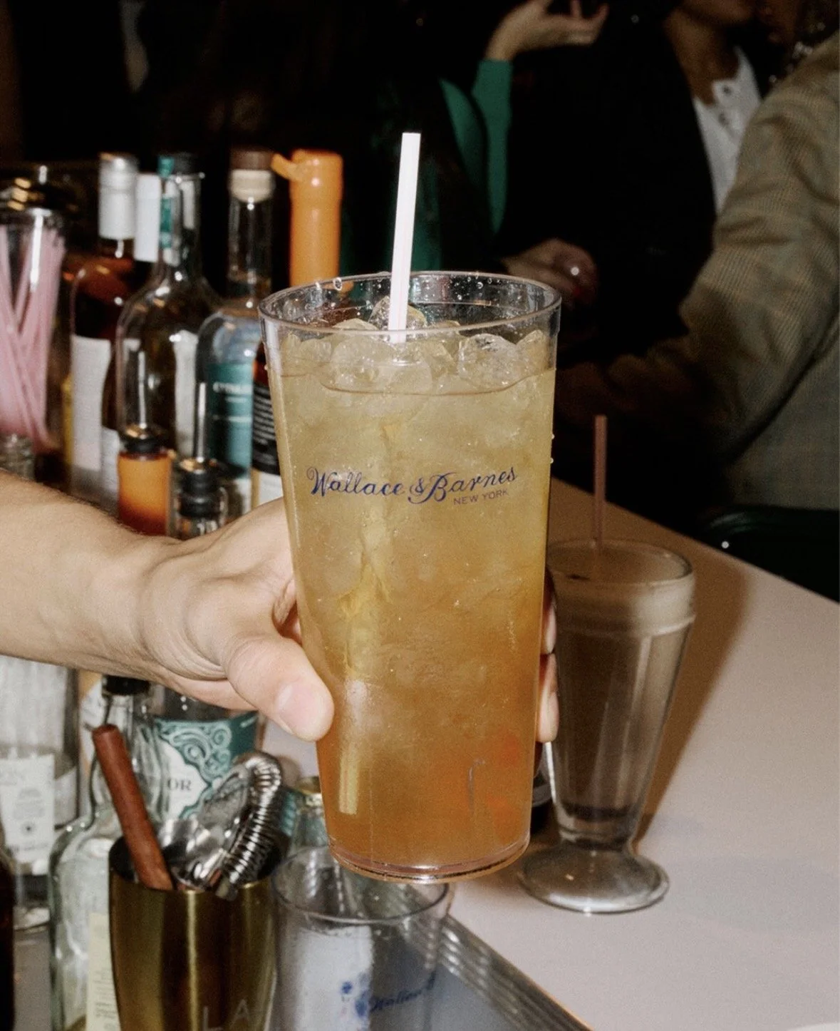 A hand holding a tall glass of iced tea with ice cubes and a straw, branded with 'Wallace & Barnes New York,' in a bar setting with bottles of alcohol in the background.