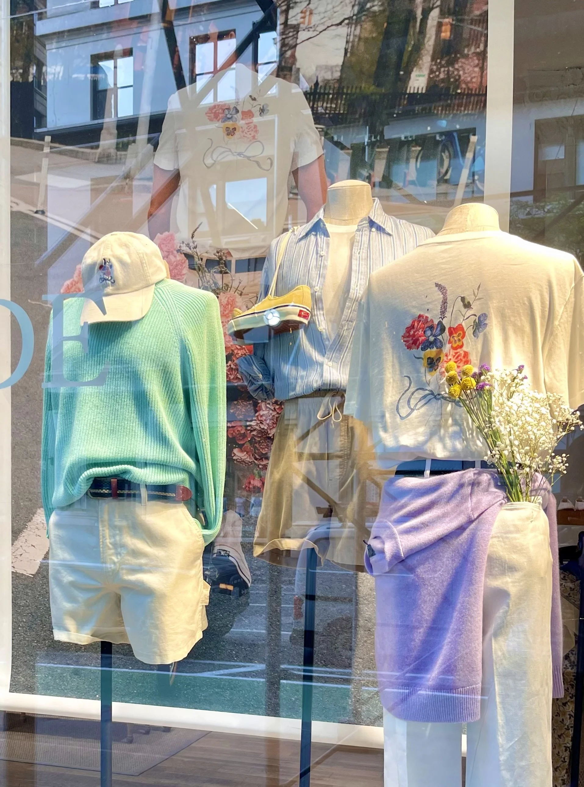 Store window display featuring mannequins dressed in casual summer clothing, including a striped shirt, pastel shorts, a pastel sweater, a cap, and a bunch of flowers.