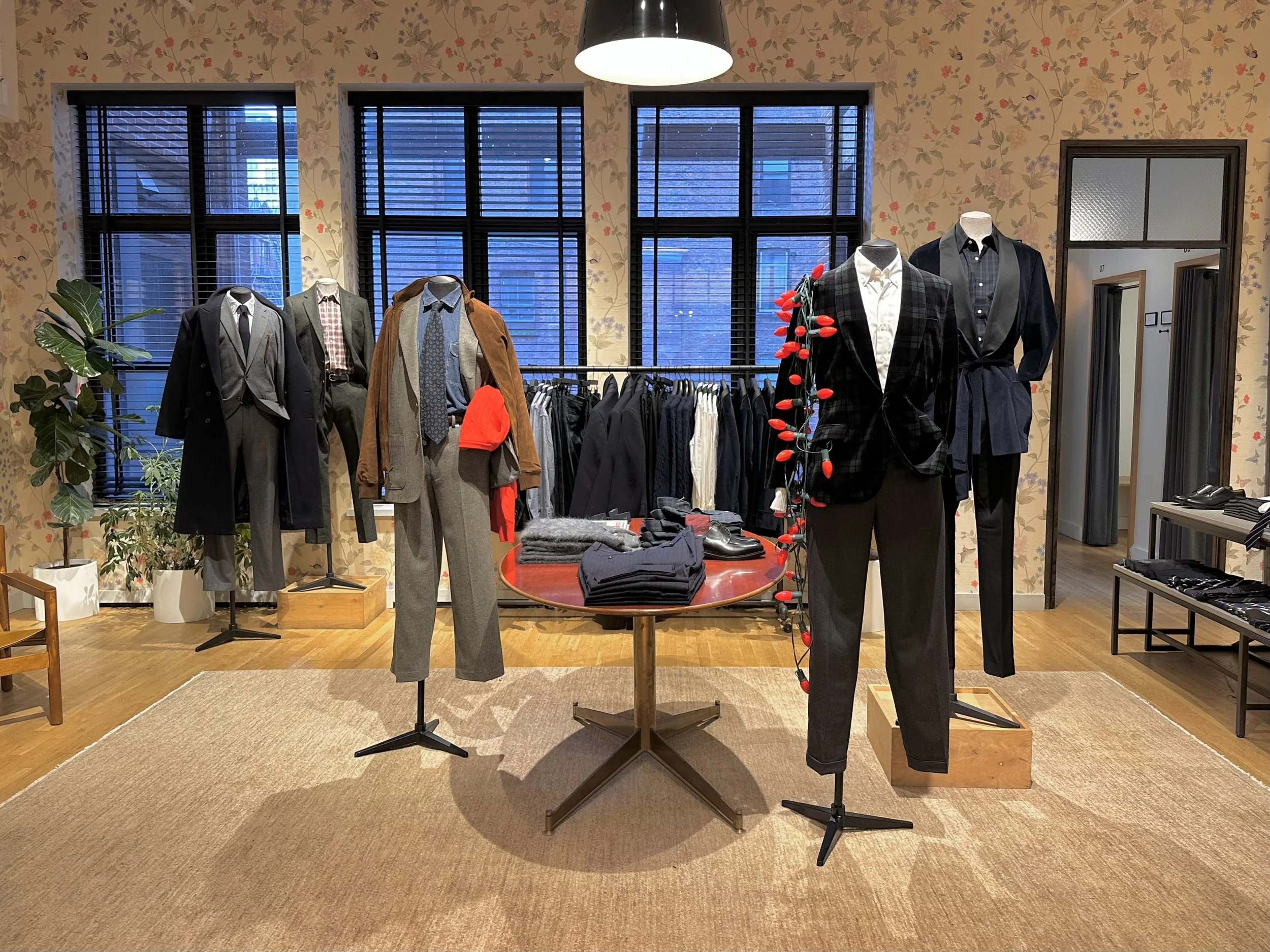 Display of men's fashion clothing including suits, trousers, jackets, and shirts on mannequins and racks inside a store with floral wallpaper and large windows.
