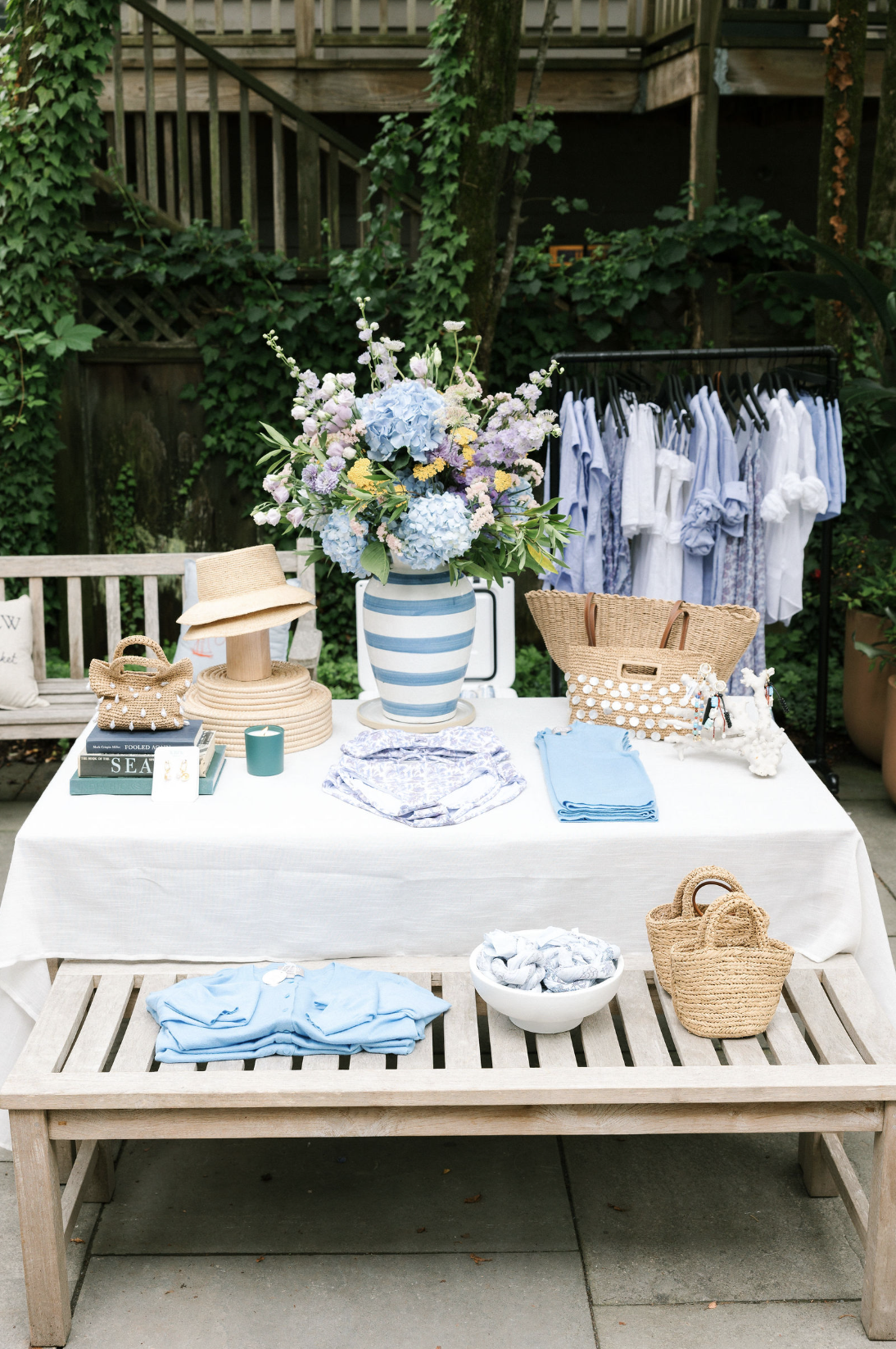 A display table with a large blue floral arrangement in a striped vase, wicker handbags, folded blue and white clothing, decorative items including a hat, books, a candle, and jewelry. Behind, there is a clothing rack with blue and white shirts and a wooden bench with recycling or trash bags.