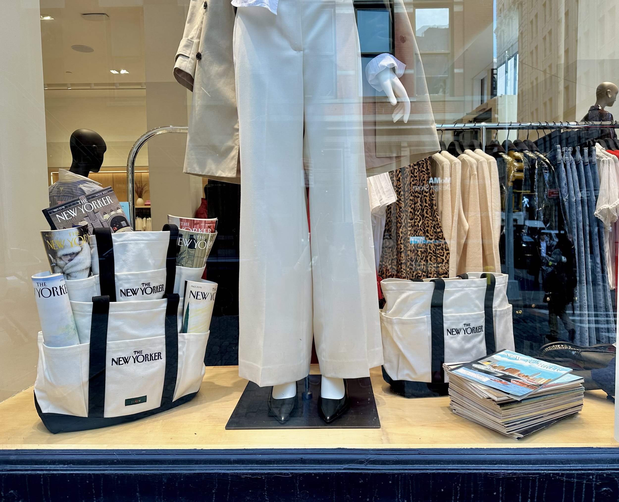 Clothing display window with mannequin dressed in beige coat and pants, blue and white gloves, and black shoes; magazines and tote bags with 'The New Yorker' logo are on the table, along with a stack of magazines.