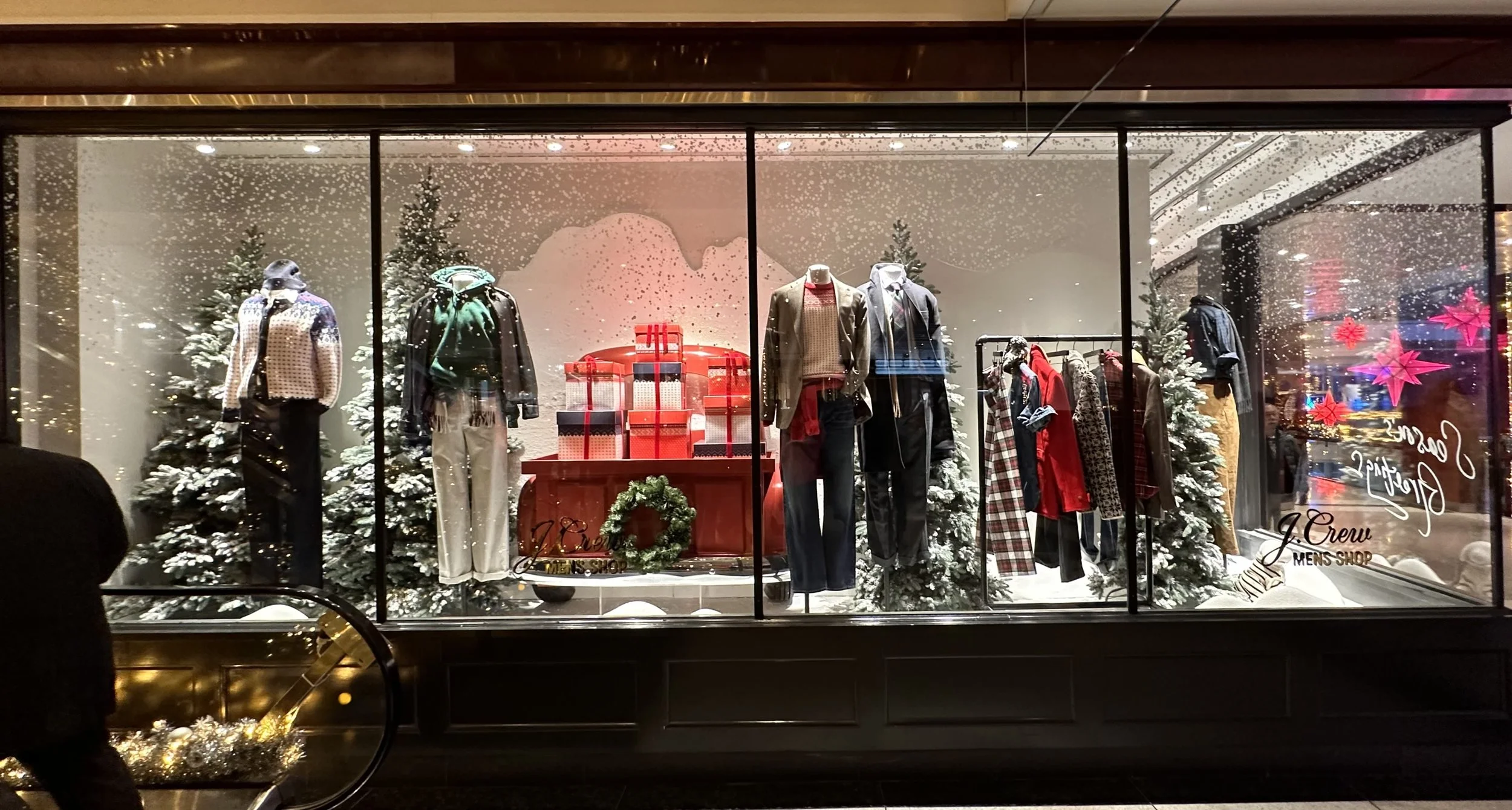 Display window decorated for Christmas with mannequins dressed in winter clothing, Christmas trees with snow, and wrapped gifts, with snow falling outside.