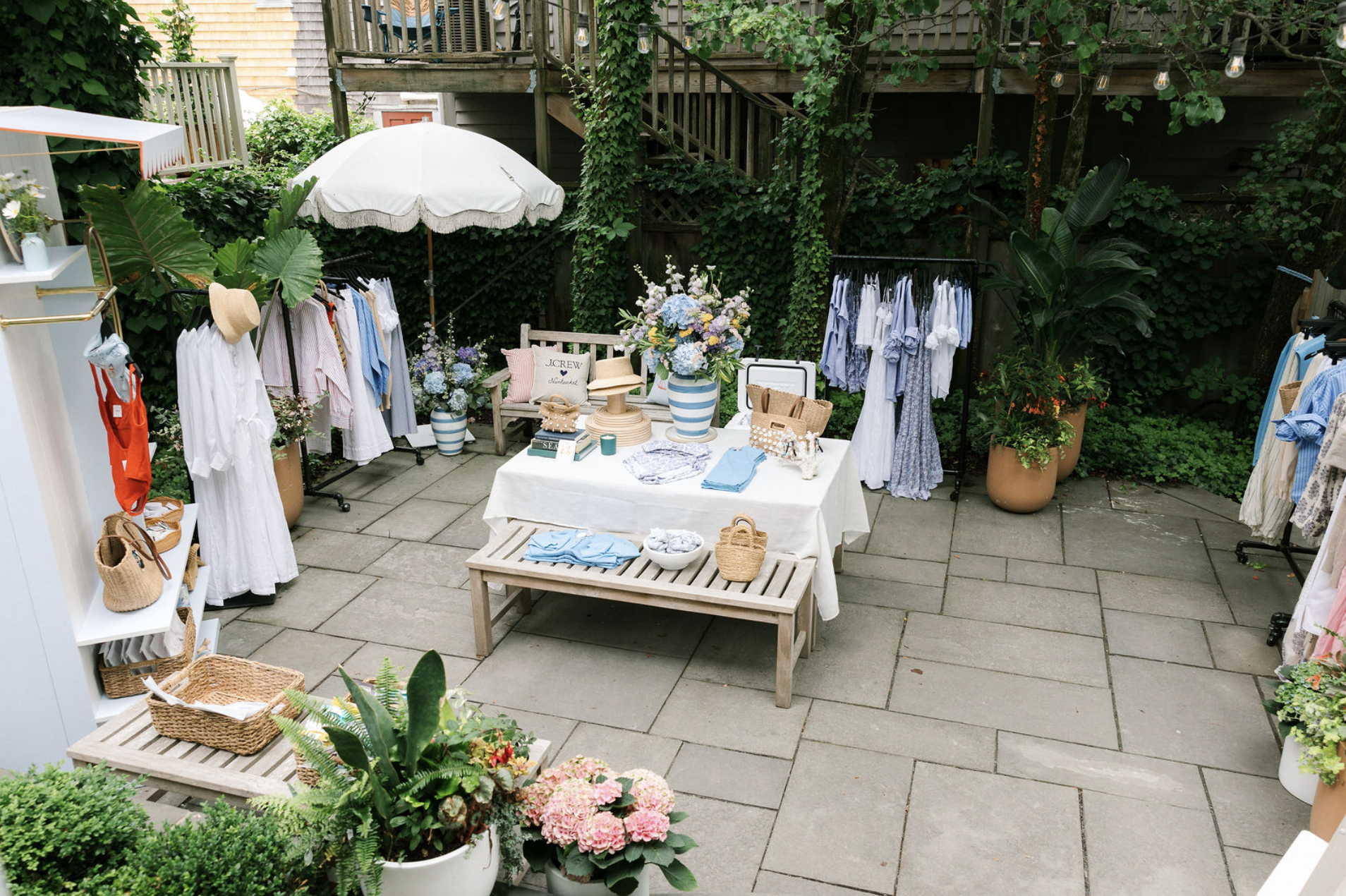 Outdoor boutique setup with clothing, accessories, and decorative items on display, surrounded by lush greenery and potted plants.