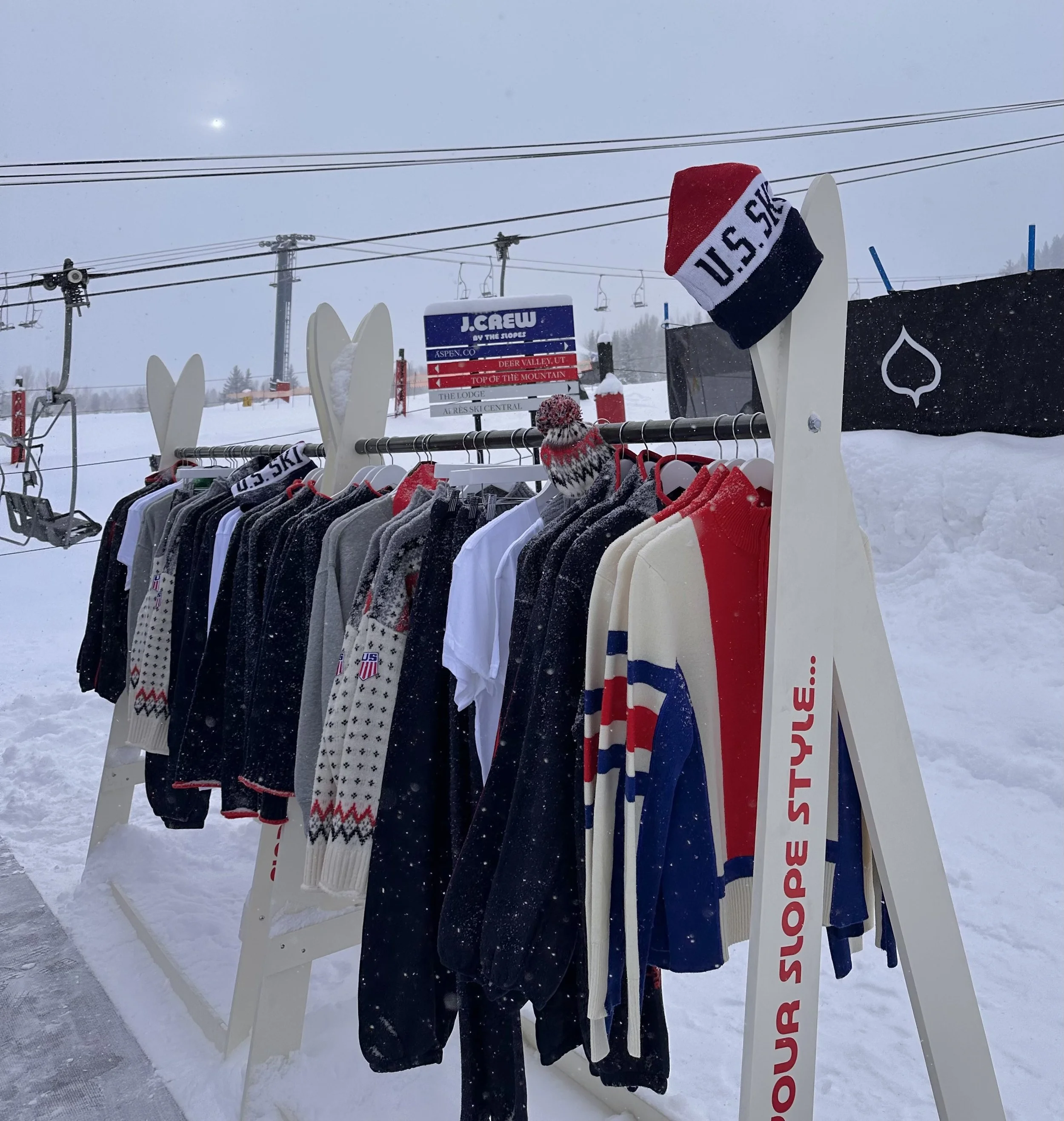 Clothing rack with ski sweaters and shirts near a ski lift at a snowy ski resort.
