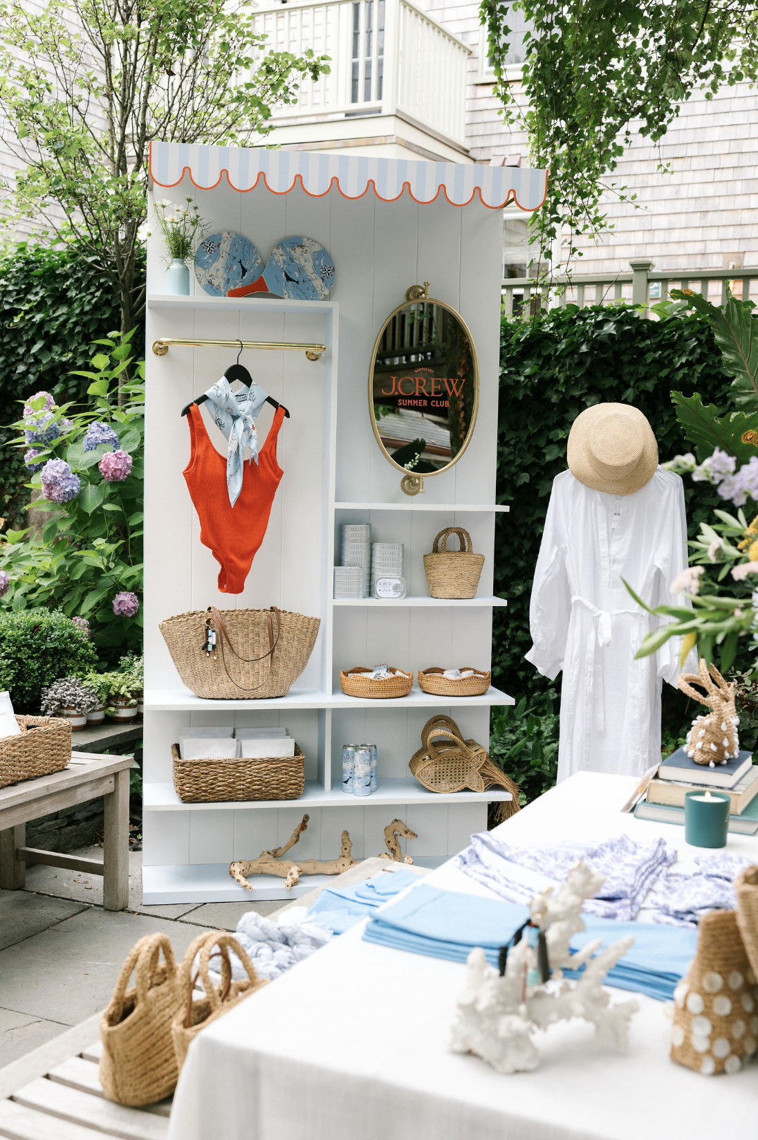 Outdoor display stand with summer clothing and accessories, including a white dress, straw hat, woven bags, and decorative items, set in a garden.