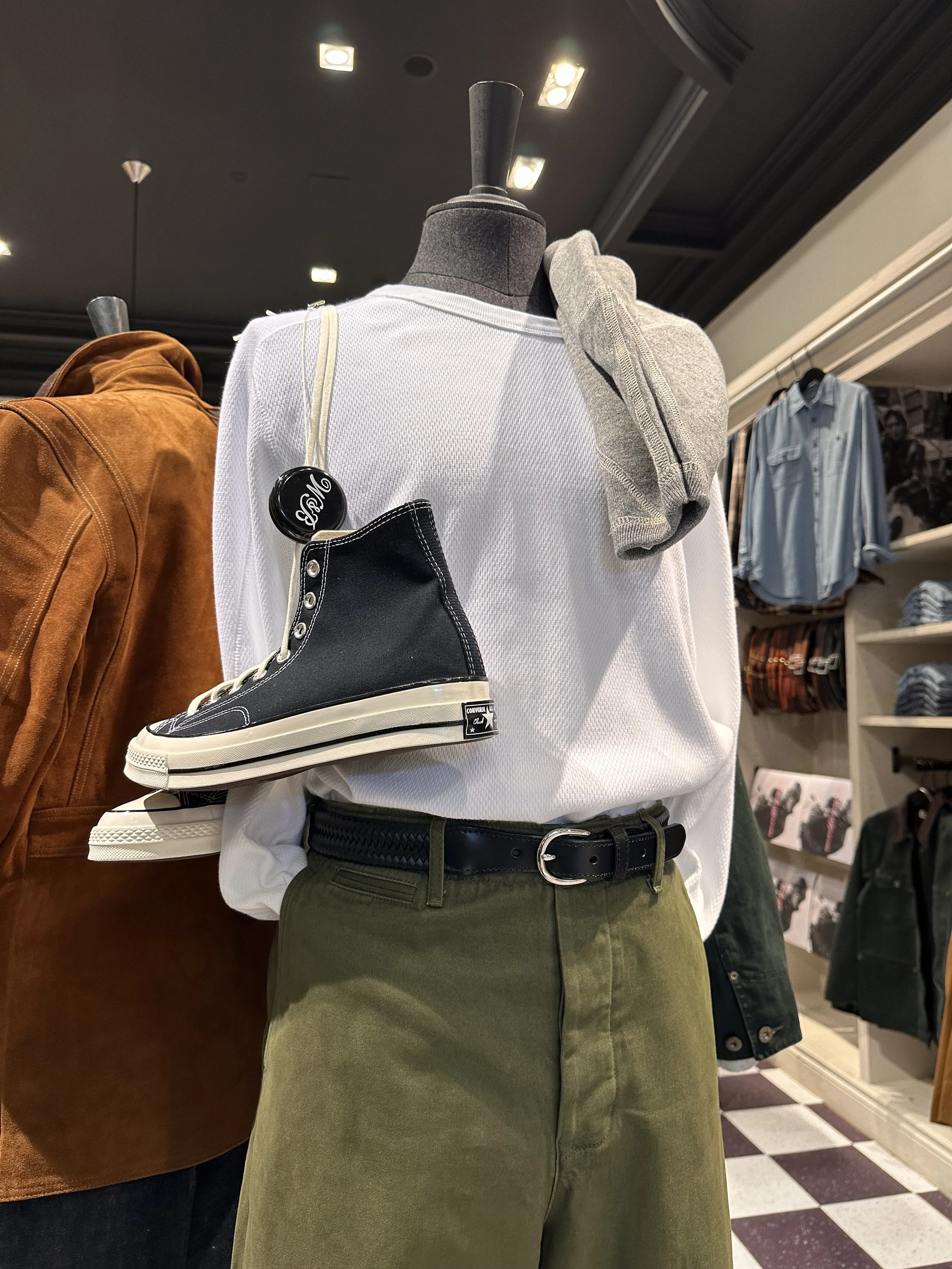 A mannequin displaying a casual outfit in a clothing store, featuring an olive green skirt, a black belt, a white sweatshirt, a gray hoodie draped over the shoulder, and a pair of black high-top Converse sneakers tucked under the sleeve.
