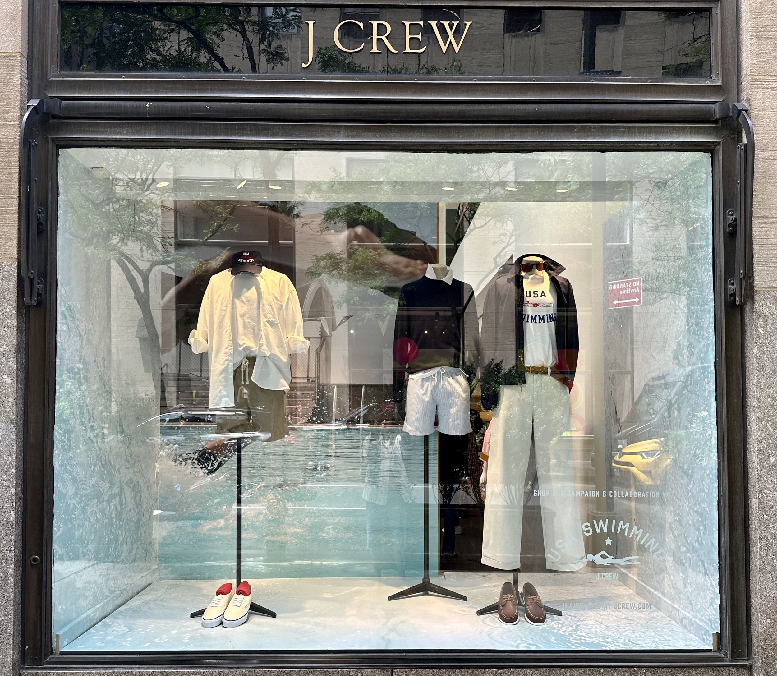 Fashion mannequins in a J.Crew storefront window display wearing casual summer outfits, including shirts, shorts, and accessories.