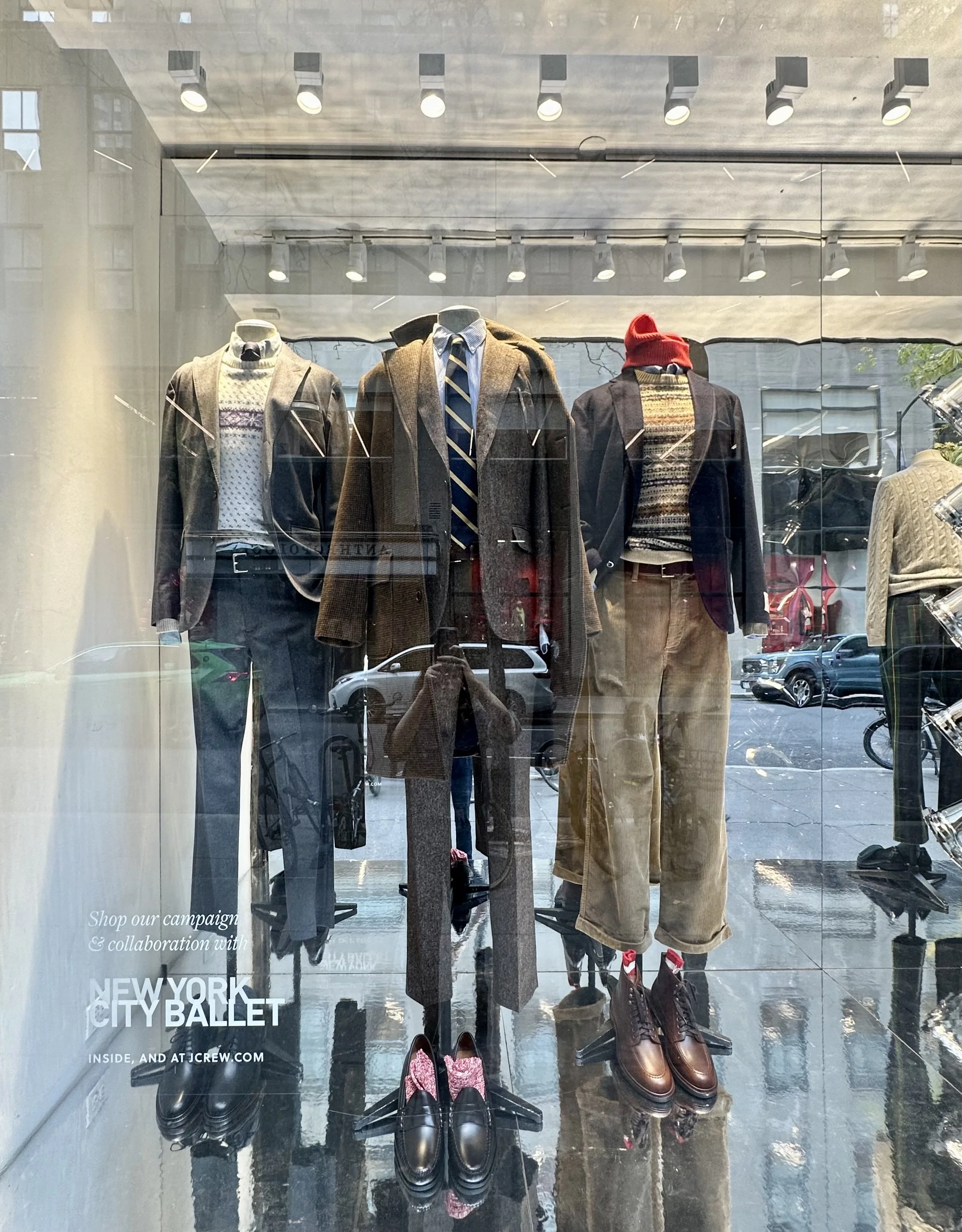 Display window featuring mannequins dressed in men's autumn fashion, including blazers, trousers, a sweater, and accessories like shoes and a scarf, with reflections of the street outside.