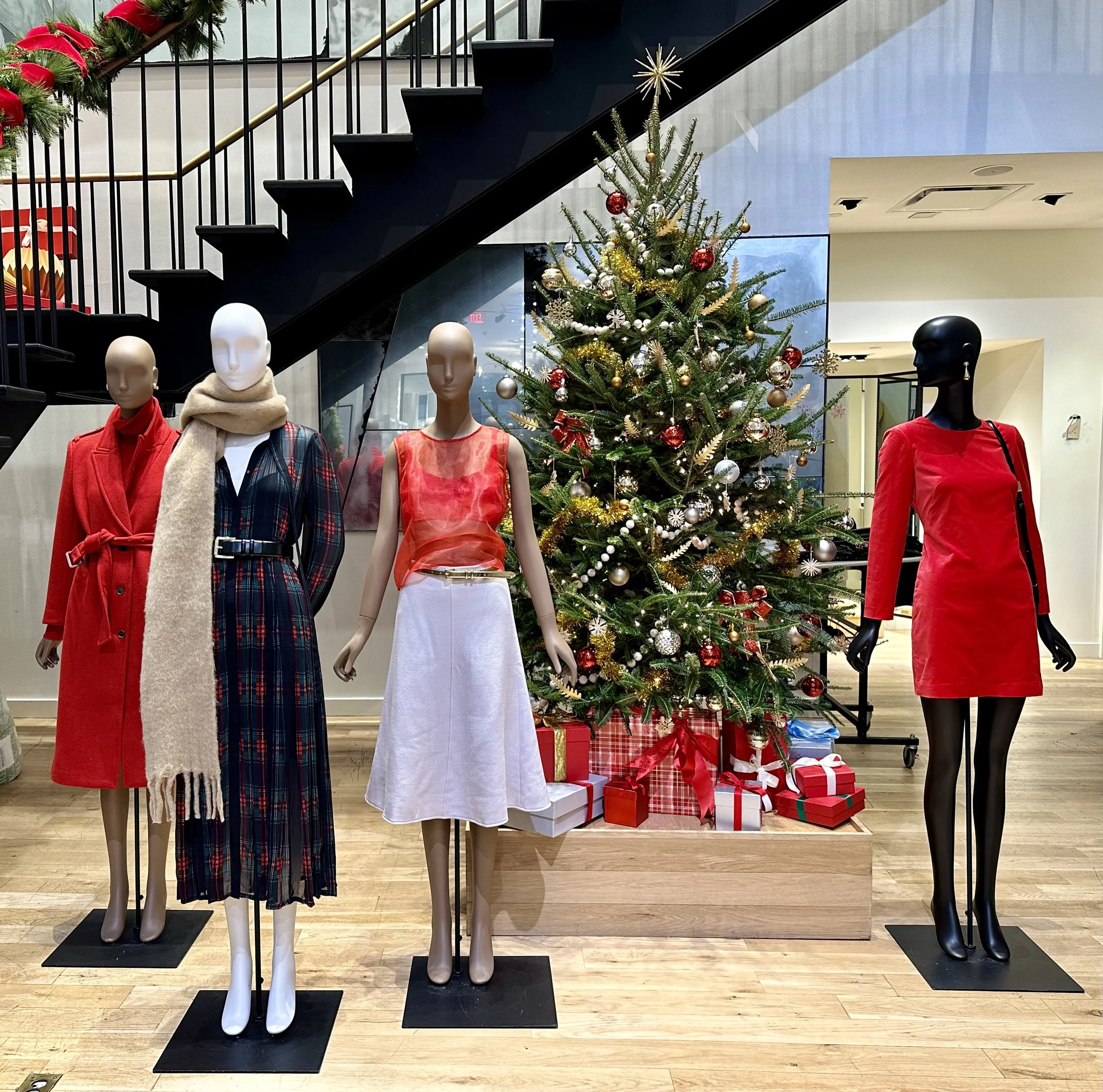 Display of mannequins dressed in holiday fashion in front of a decorated Christmas tree with presents underneath, inside a store with a wooden floor and a staircase.