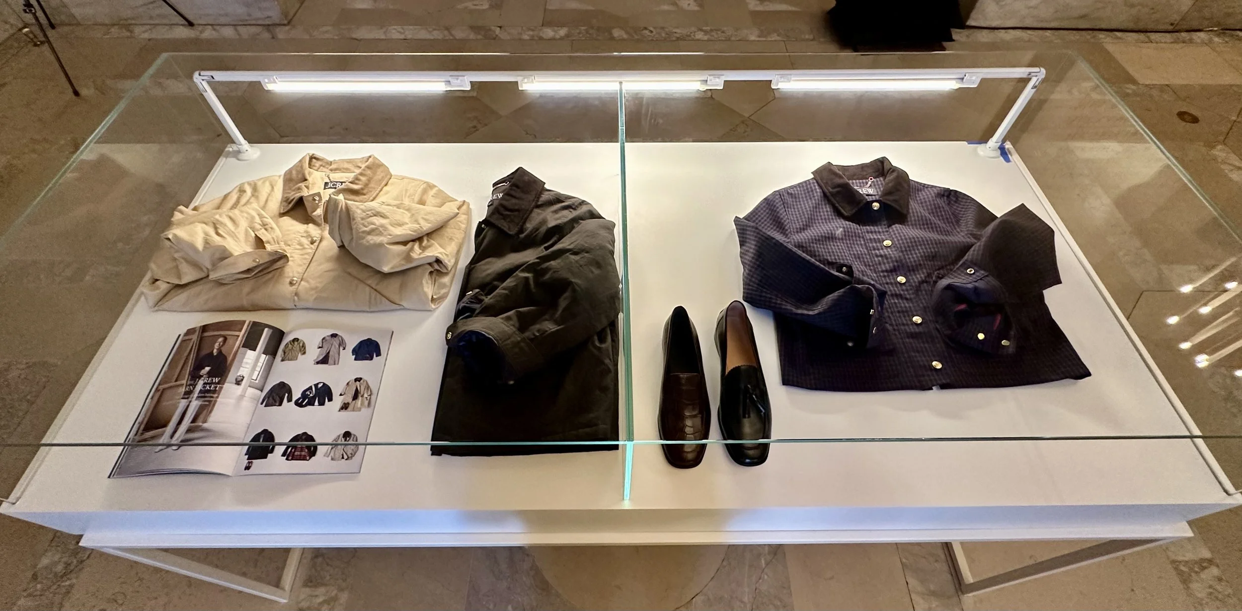 Display of men's clothing and shoes in a glass case, including shirts, folded pants, shoes, and a fashion catalog.