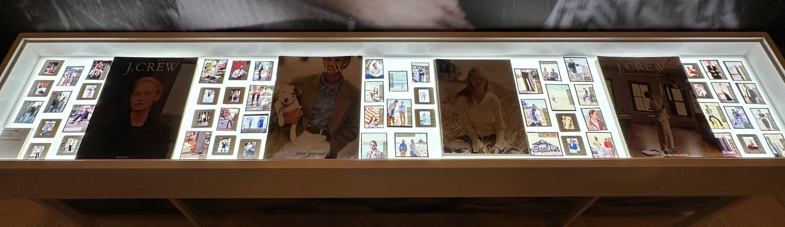 Backlit display case with photographs and advertisements, including J.Crew posters and various fashion images.