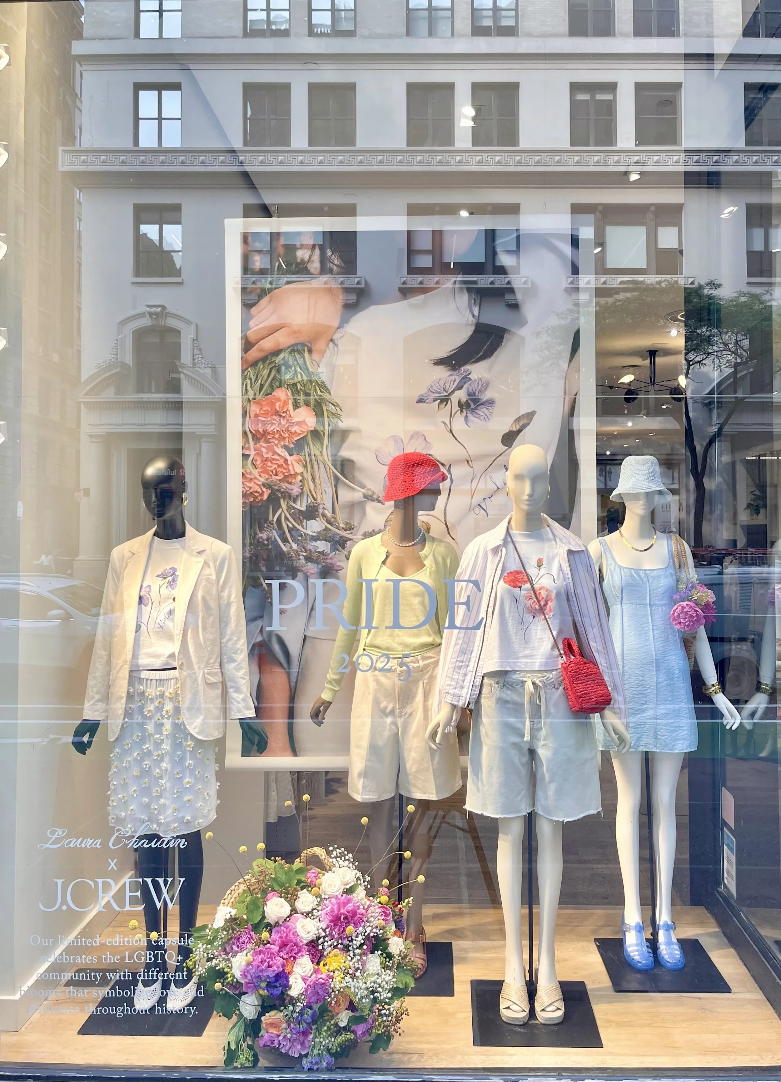 Display window with mannequins dressed in white and pastel clothing, behind a large poster with flowers, and floral arrangements in front. The display promotes Pride 2023.