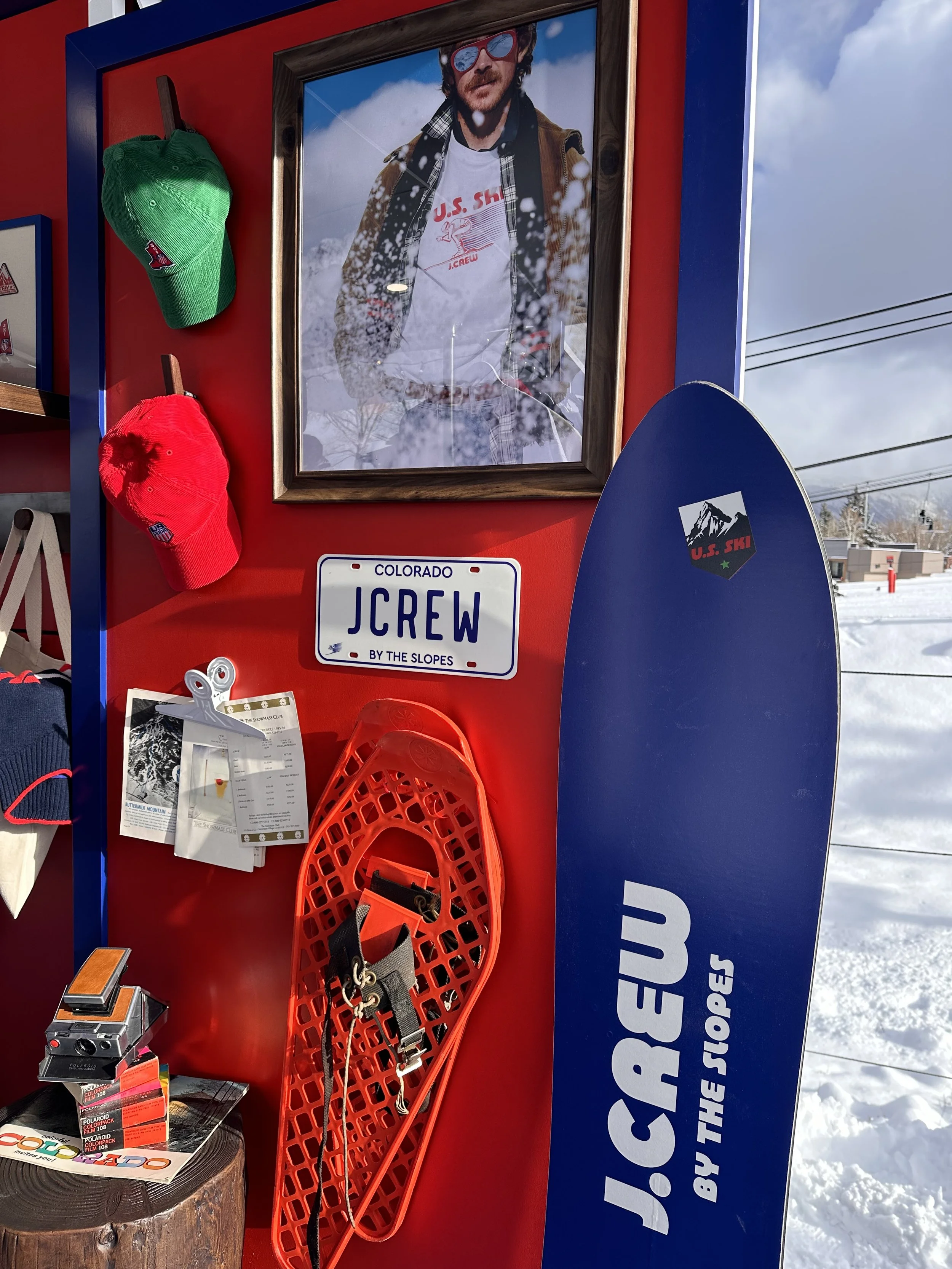 Photo of a display wall with a large framed picture of a man in winter clothing, a green and red ski hat, a snowboard, a Colorado license plate with 'JCREW BY THE SLOPES,' and snow outside.