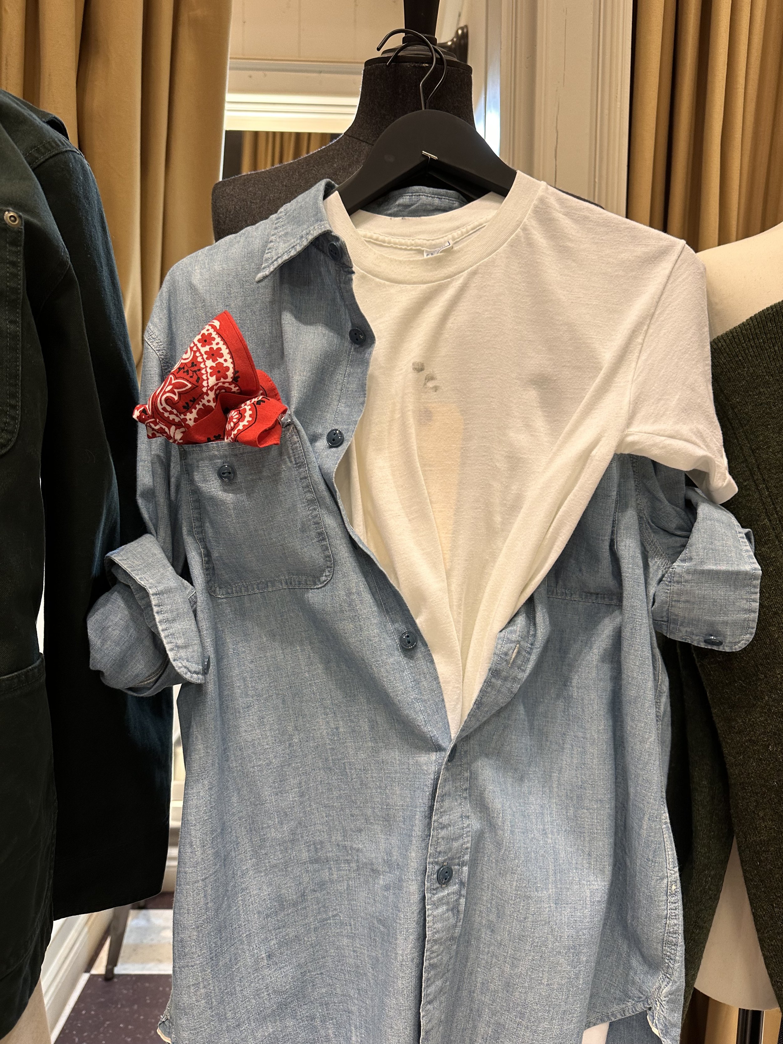 A mannequin wearing a light blue denim shirt over a white t-shirt with a red bandana tucked into the shirt's pocket, on display on a clothing rack.