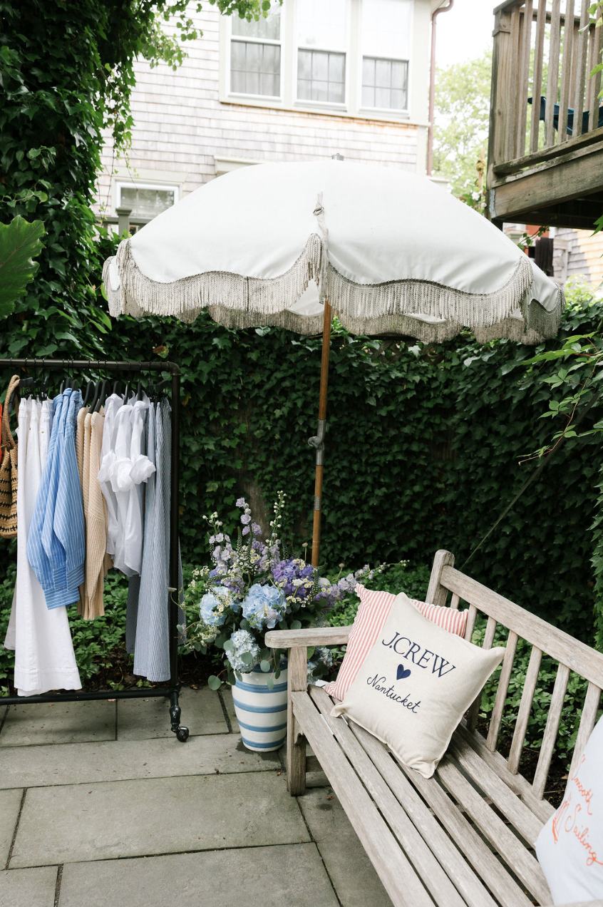 A cozy outdoor patio corner with a white umbrella, striped bench with decorative pillows, a metal clothing rack with hanging clothes, and a large blue and white striped flower pot with purple and white flowers, all surrounded by lush green ivy.