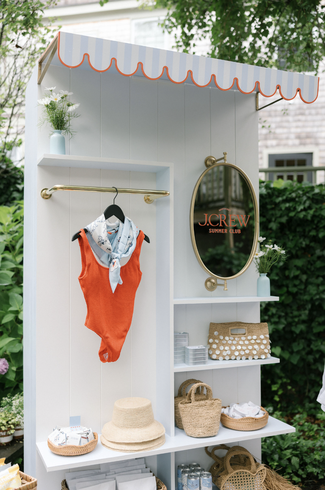 A white outdoor display stand with a red and white striped canopy, featuring a mirror with the J.Crew Summer Club logo, a small vase with white flowers, a basket with folded textile items, woven baskets, a stack of cans, and a red and white sleeveless top with a scarf draped over it.