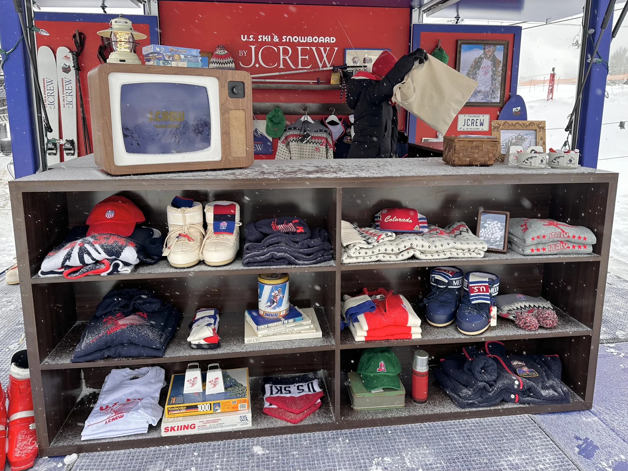 Display of U.S. ski and snowboard accessories and apparel at a ski resort, including hats, gloves, socks, shoes, mugs, and framed pictures, with snow on the ground and a person in winter clothing inside the booth.