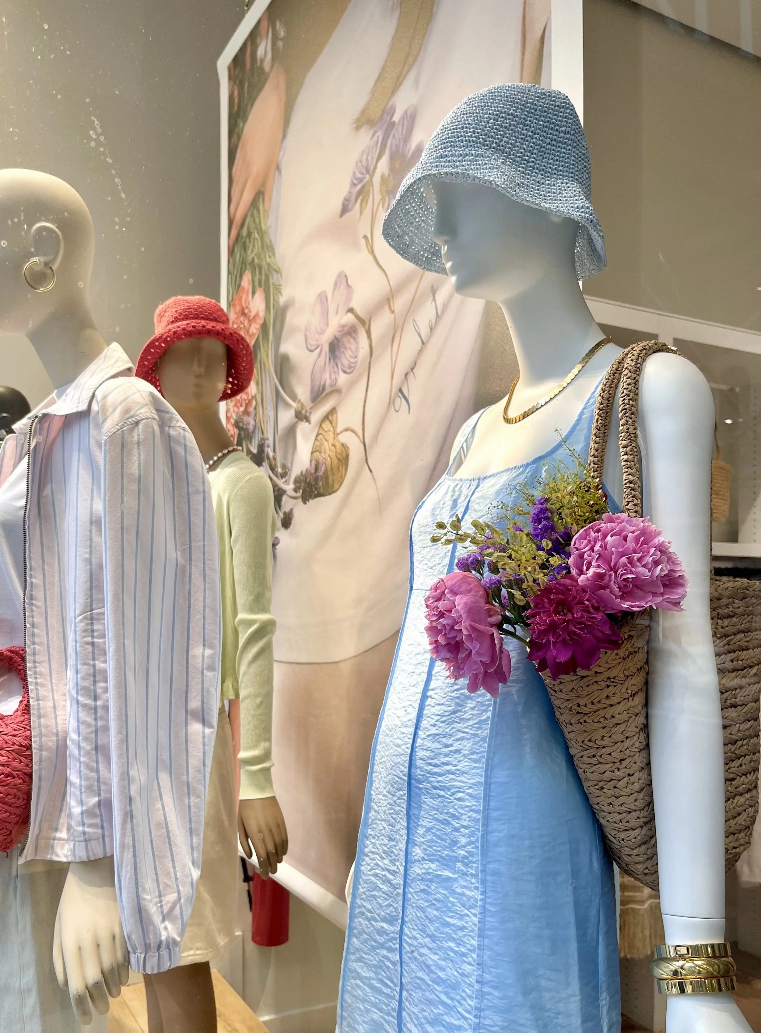 Display of mannequins dressed in summer clothing, with accessories like hats and jewelry, in a store window. One mannequin wears a blue dress, tan bag with flowers, and a light blue hat. Another has a yellow top, pink hat, and striped oversized shirt layered over it. A poster with floral embroidery hangs behind.