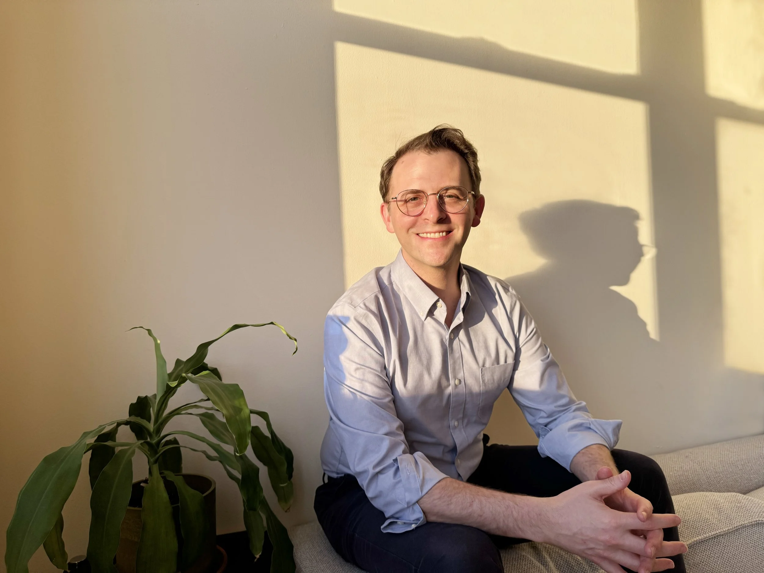 A smiling man with glasses, wearing a light-colored shirt, sitting on a beige couch next to a green potted plant, with sunlight casting shadows on the wall behind him.