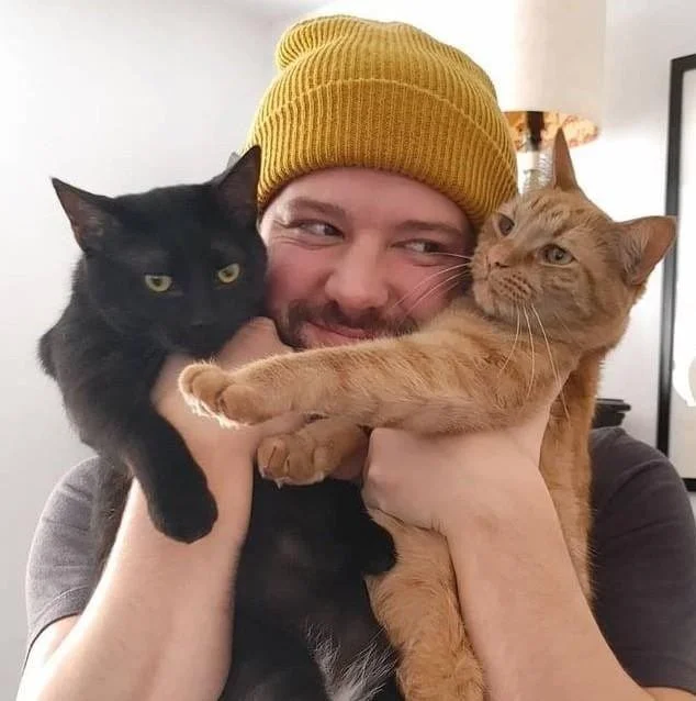 Person wearing a yellow beanie holding two cats, one black and one orange, close to their face indoors.