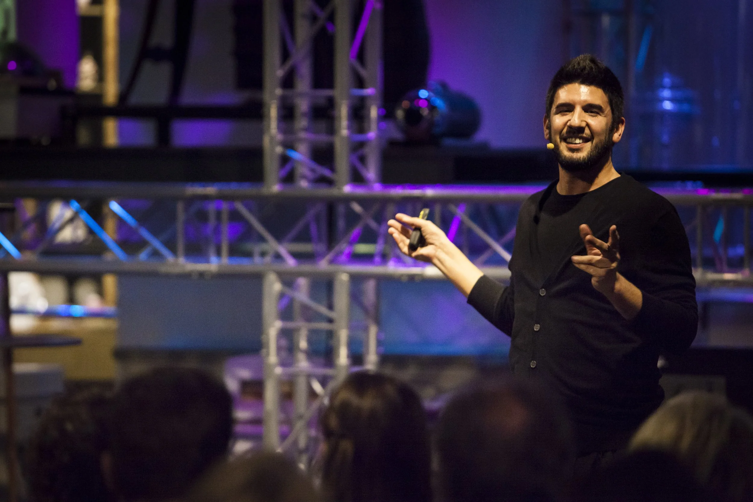 Jorge Alves Lino, a man in a black shirt speaking to an audience at a conference or event, gesturing with one hand and holding a remote in the other, with stage lighting and metal framework in the background.