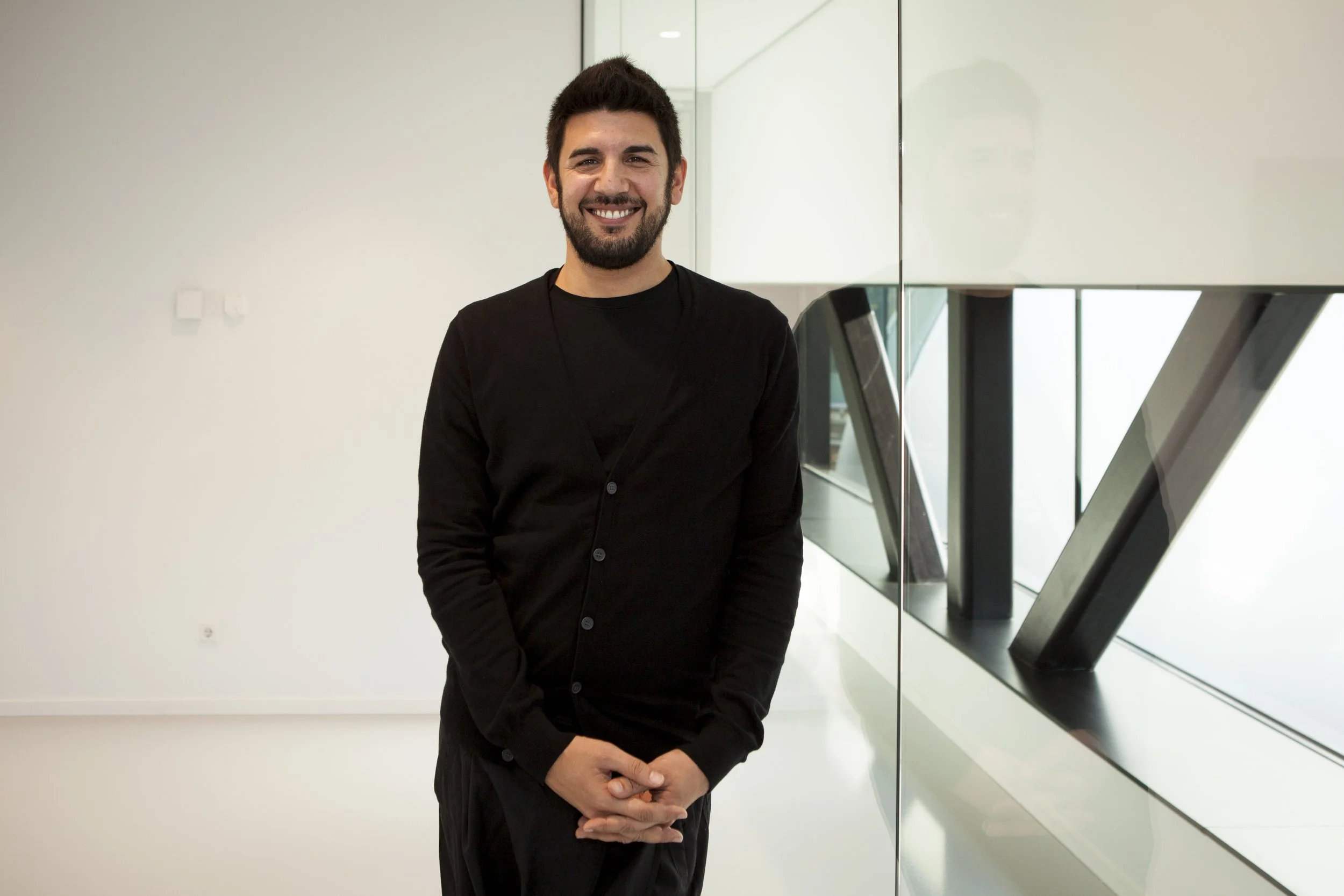 Jorge Alves Lino, a smiling man with dark hair and a beard, wearing a black shirt, standing indoors near a glass wall with windows.