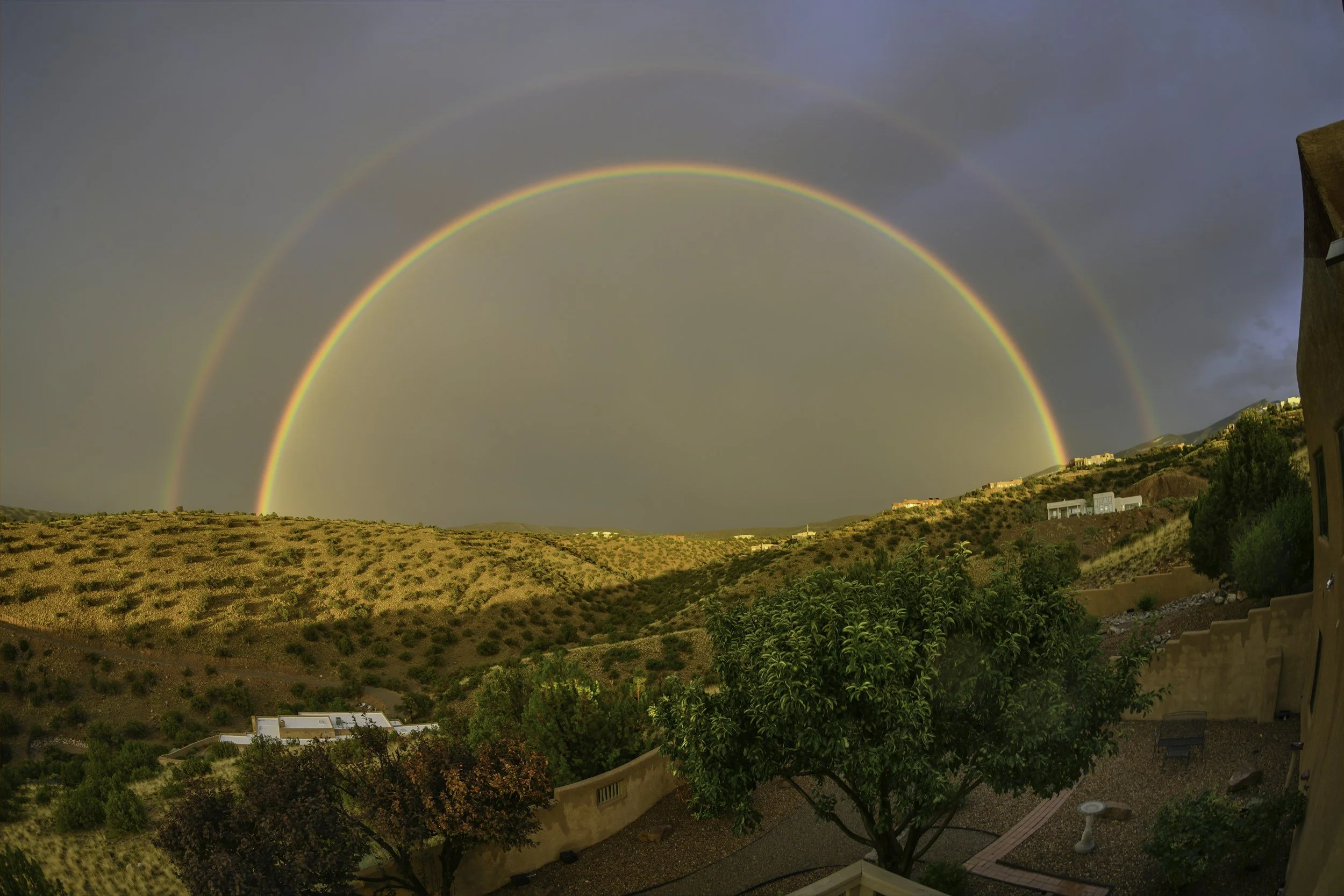 A double rainbow over a desert landscape with hills, houses, and trees during a storm.