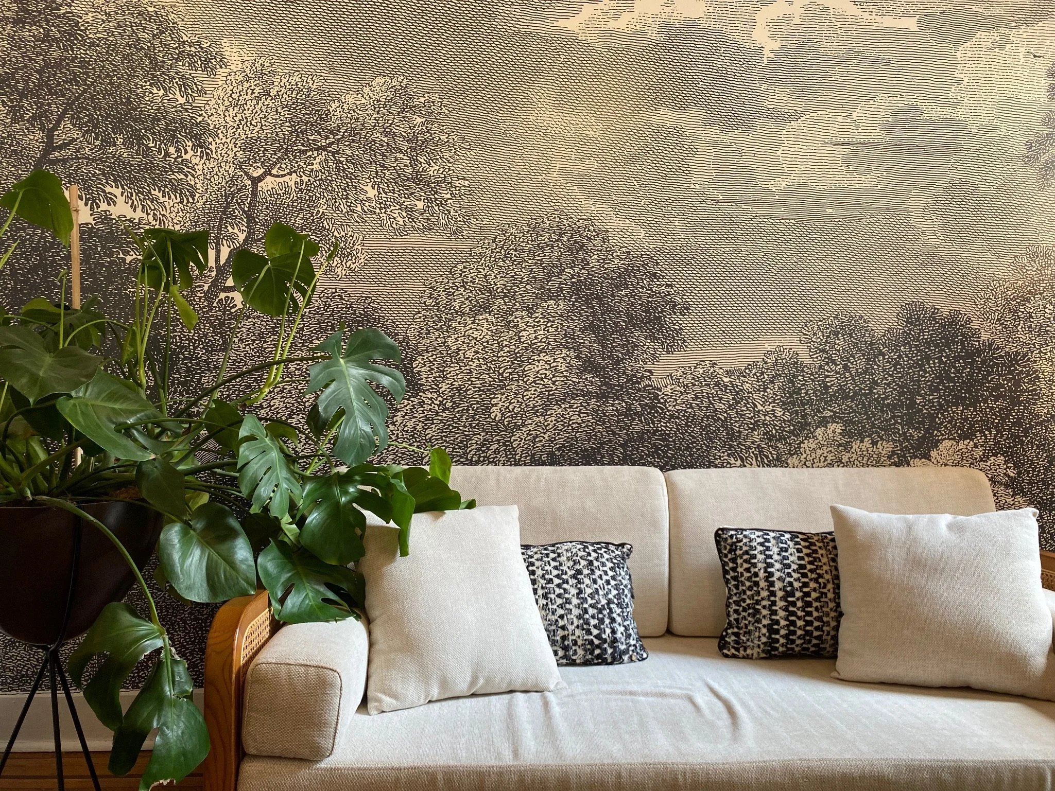 Living room with a beige sofa, patterned throw pillows, a tall potted Monstera plant, and a decorative wall mural of trees and landscape scenes in black and beige.