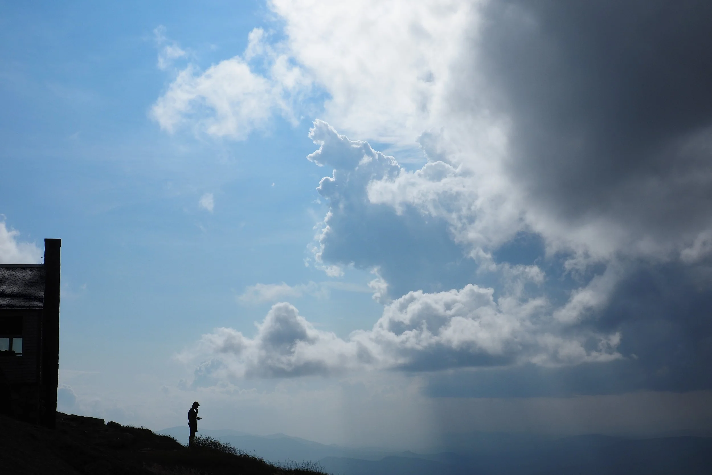 Silhouette of a person standing on a hill with a house nearby, looking at a cloudy sky with sunlight breaking through.