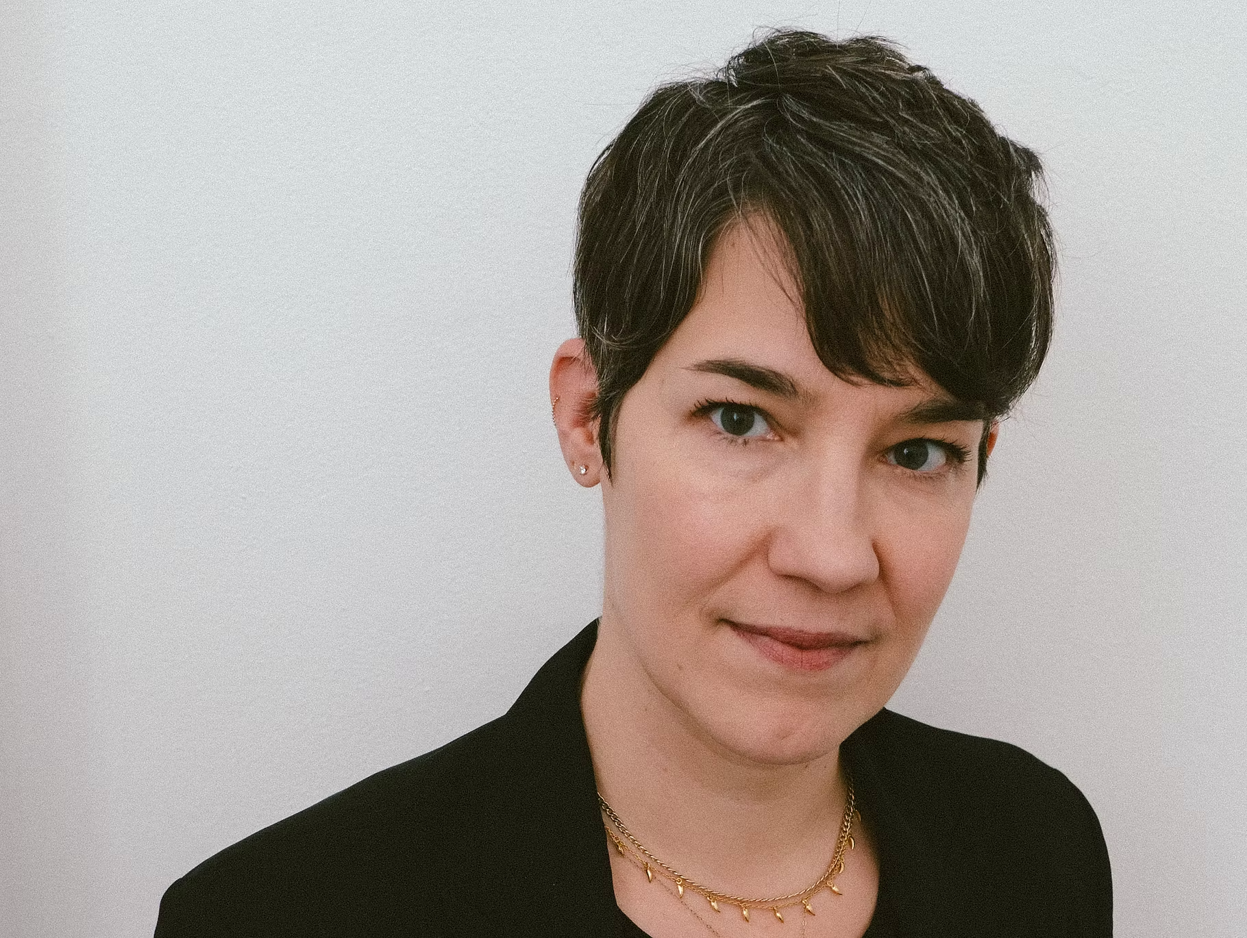 Portrait of a woman with short dark hair, wearing a black blazer, gold necklaces, and small earrings, standing against a white background.