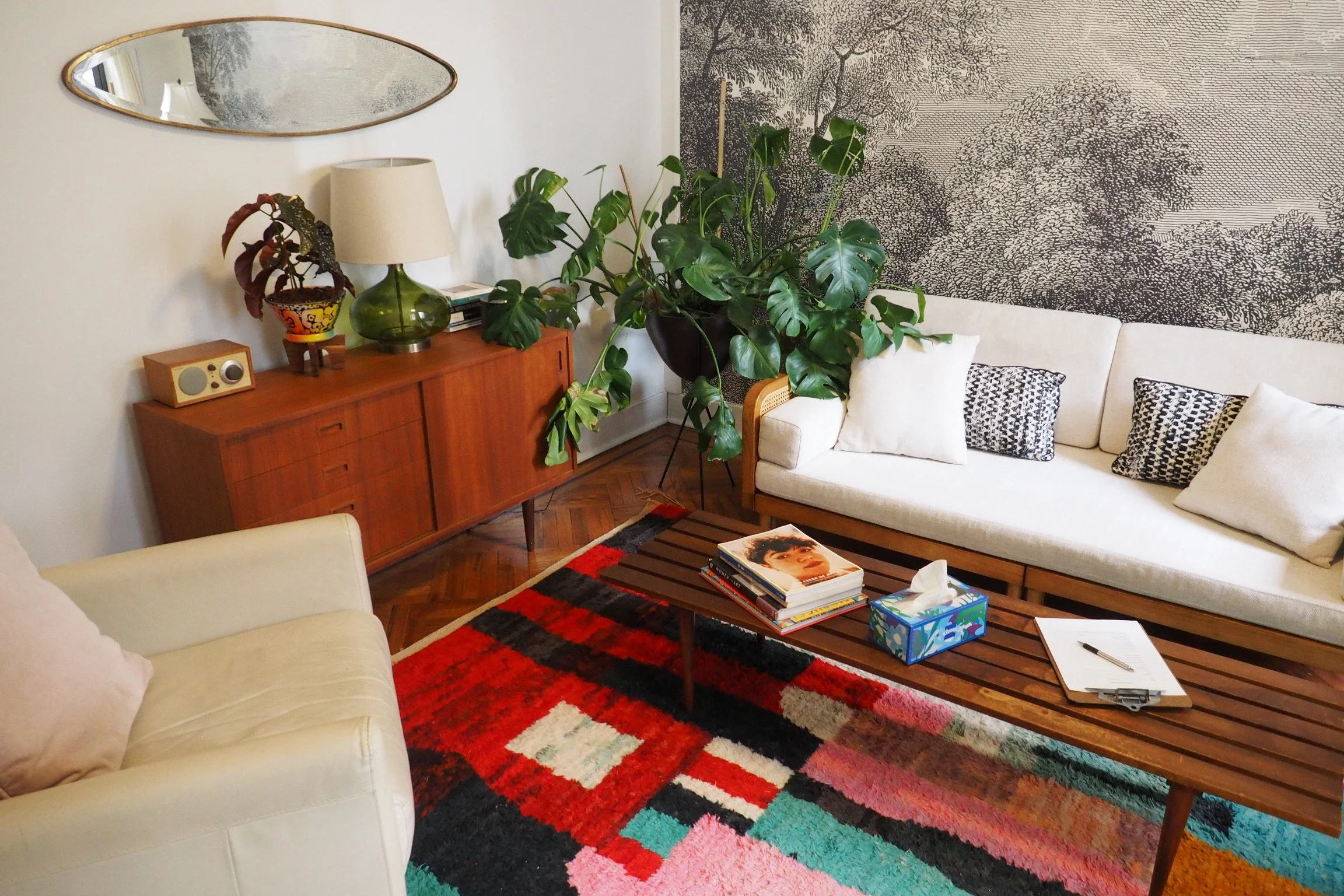 Living room with white sofa, beige armchair, wooden coffee table with magazines and tissue box, colorful geometric rug, large green houseplant, wooden sideboard with lamp, decorative items, and a mirror on the wall, with black and white patterned wal
