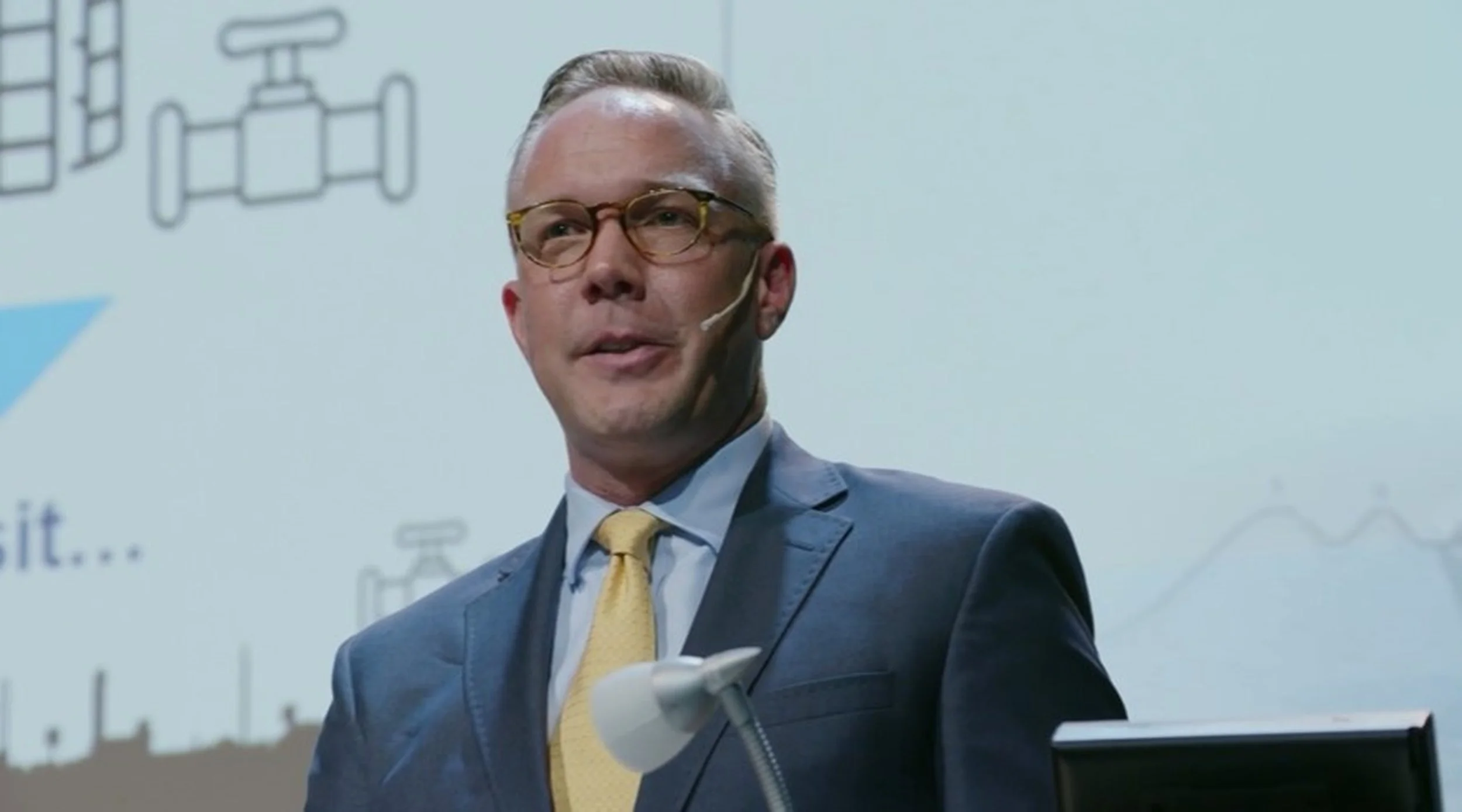 A man in a suit and yellow tie speaking at a presentation with graphics and charts on the slide behind him.