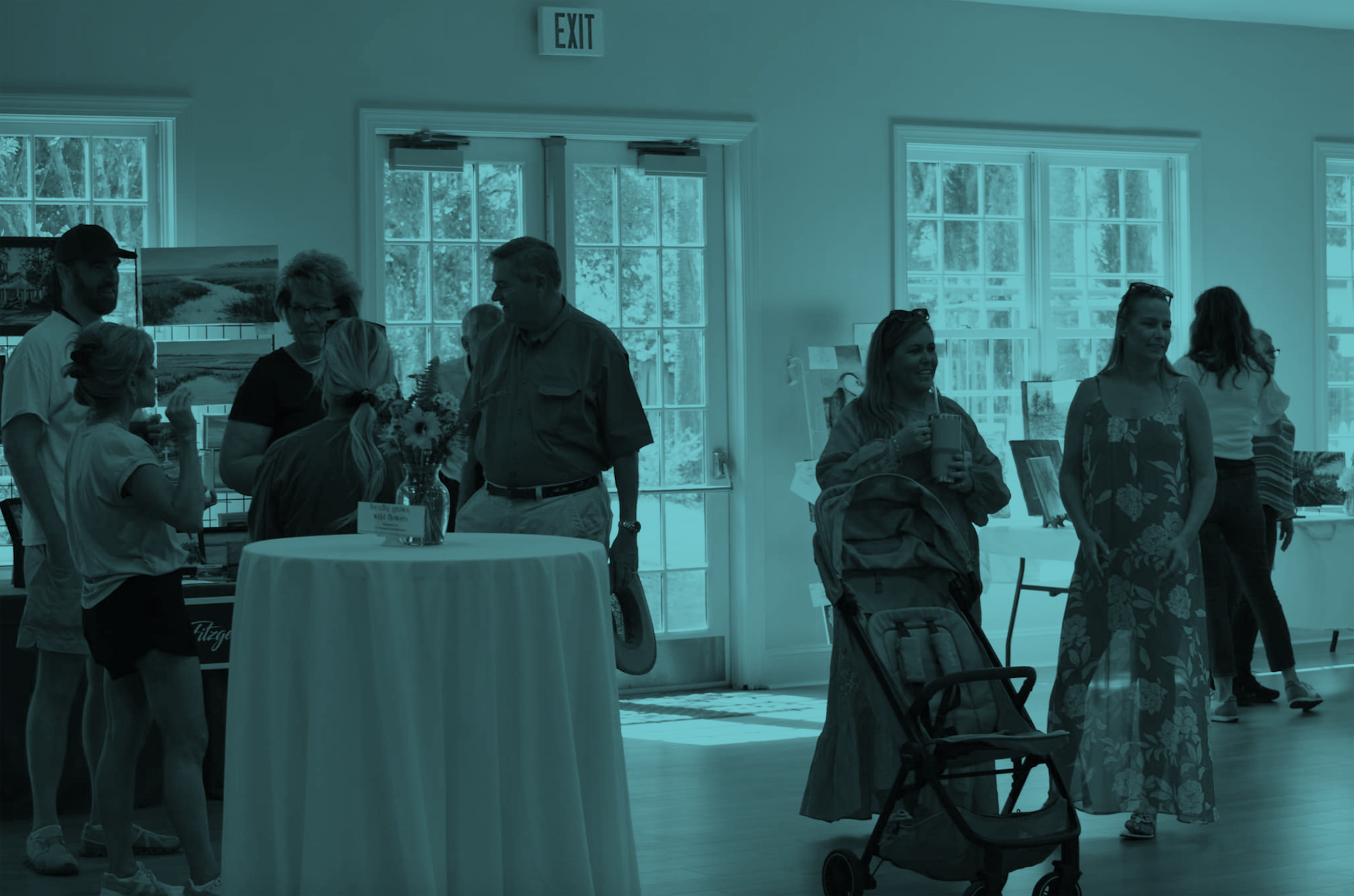 Group of people inside a bright room with large windows, some talking and others browsing artwork on display, with an exit sign above glass doors in the background.