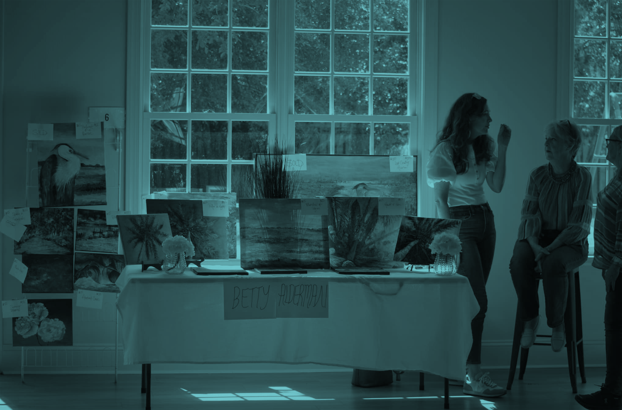 Artist Betty Alderman's art display with paintings of nature scenes on tables and walls, with four people talking inside a well-lit room with large windows.