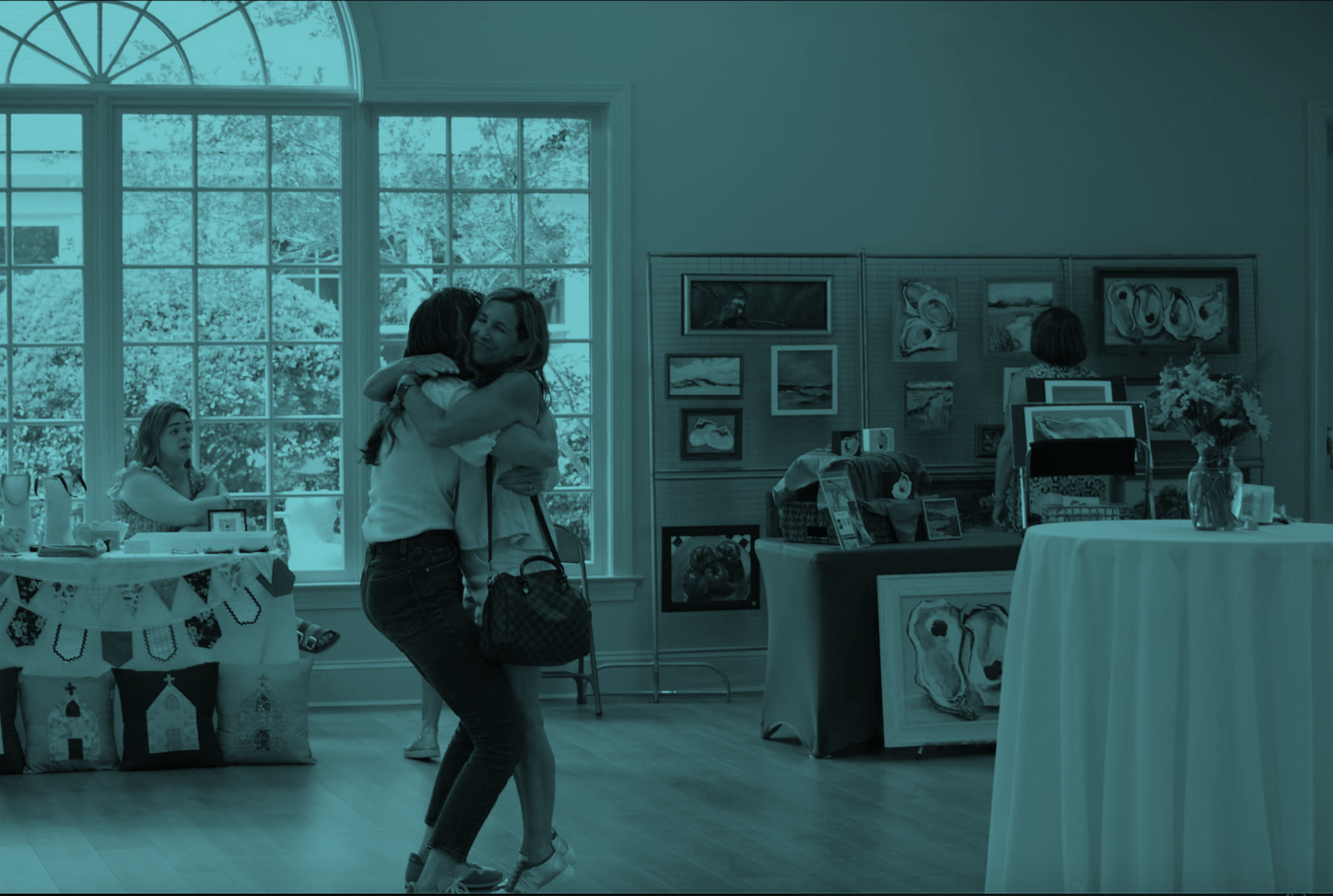 Two women hugging in an art gallery or craft fair, with a young girl sitting behind a table and another woman looking at art on display.