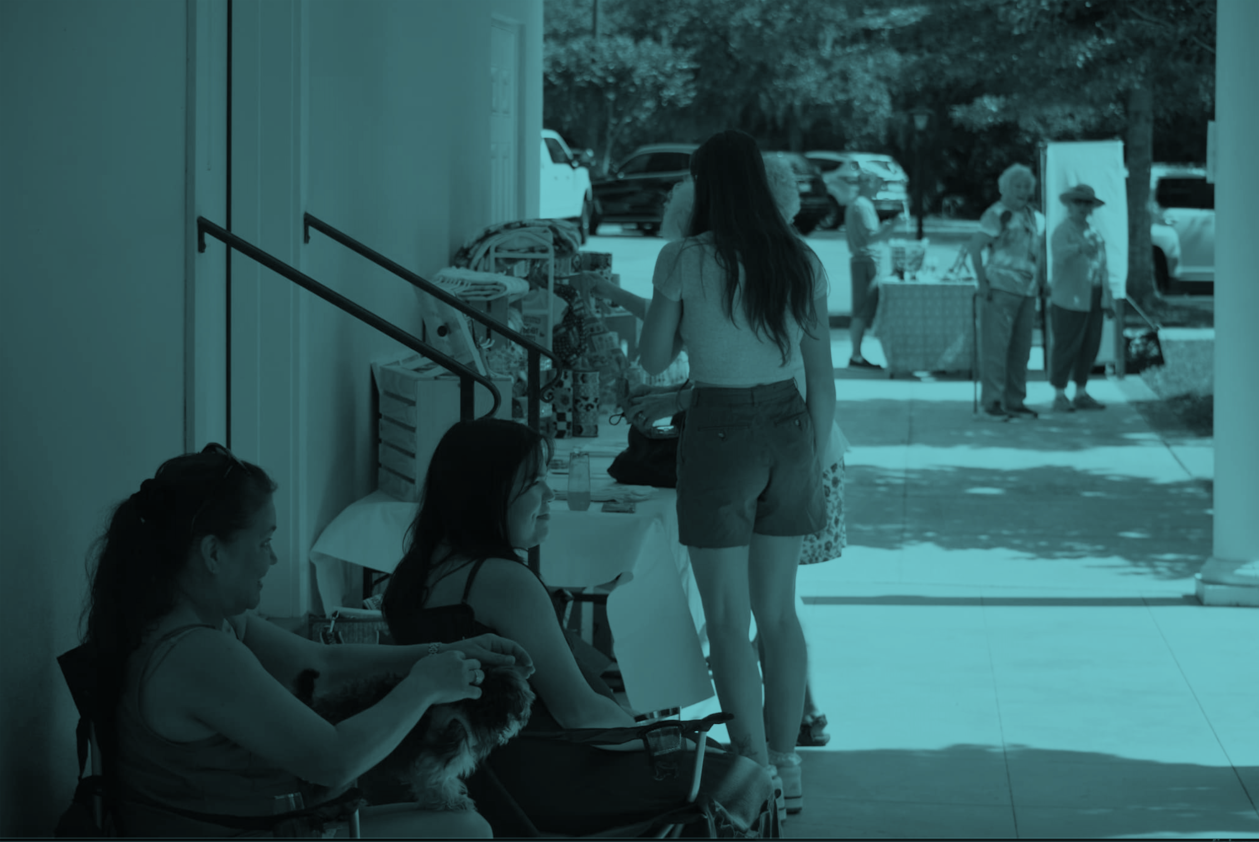 People sitting and standing outside a building with tables and chairs, some with dogs, and cars parked in the background.
