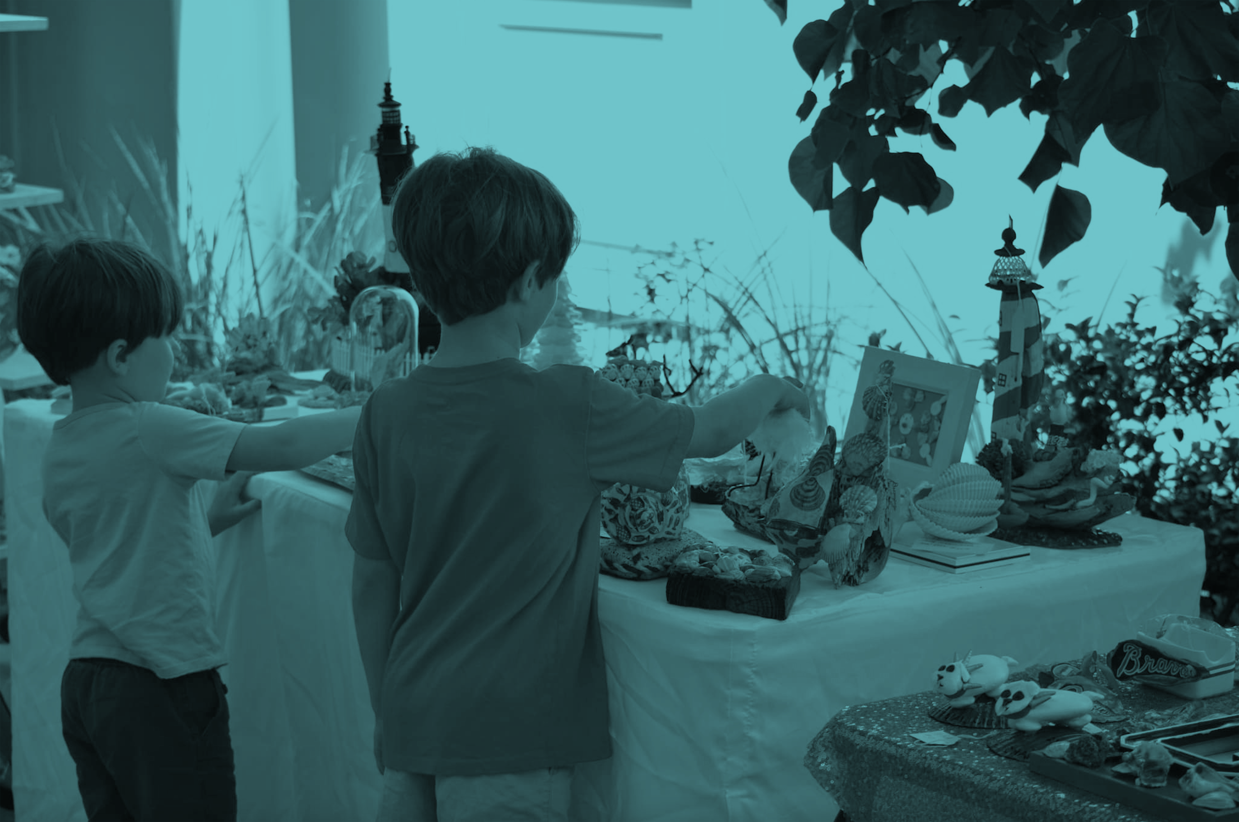 Two children shopping at a table with seashell decorations, toys, and framed pictures, in an outdoor setting with leafy plants.