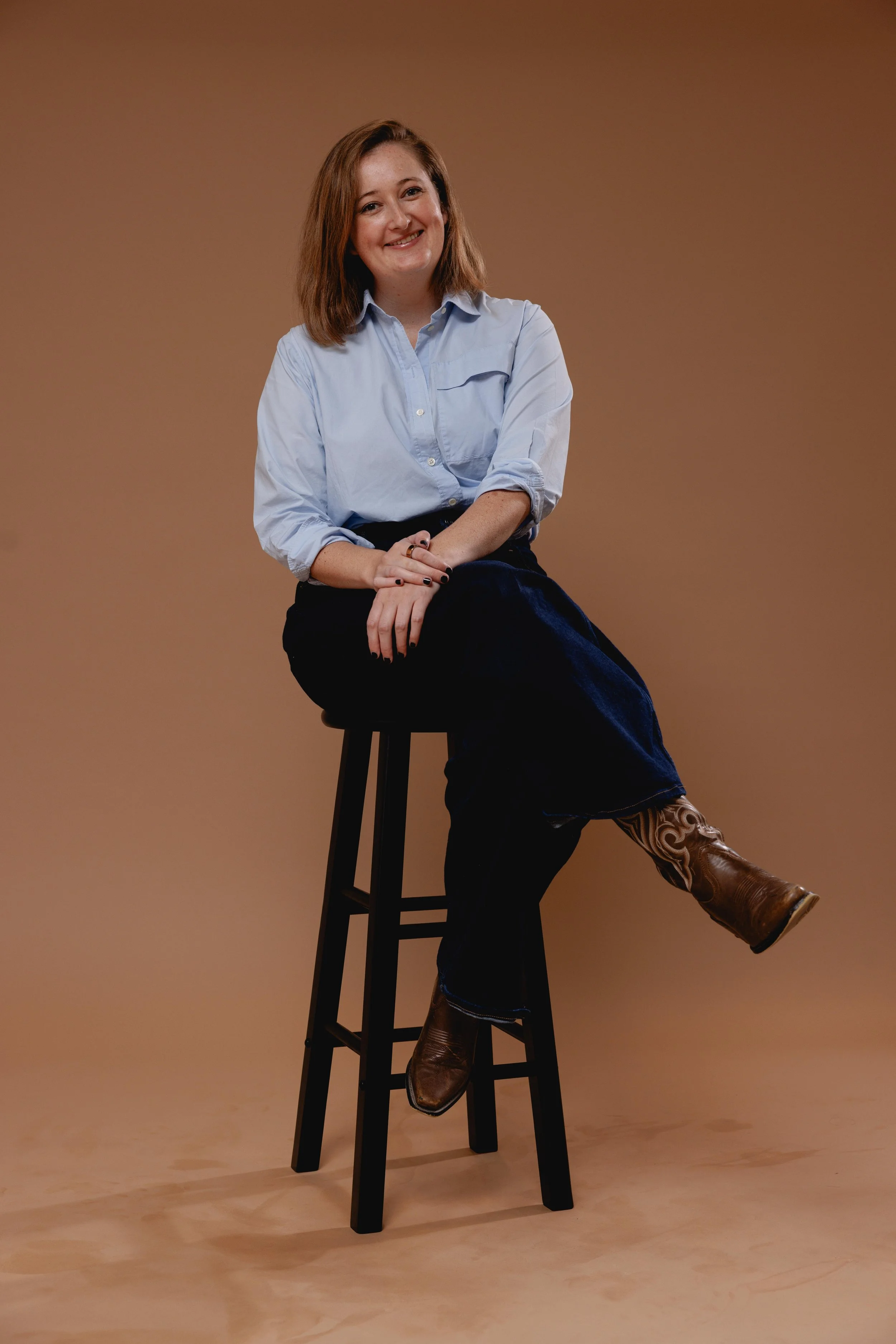 Woman sitting on a chair smiling, in a blue shirt, jeans, and brown boots.
