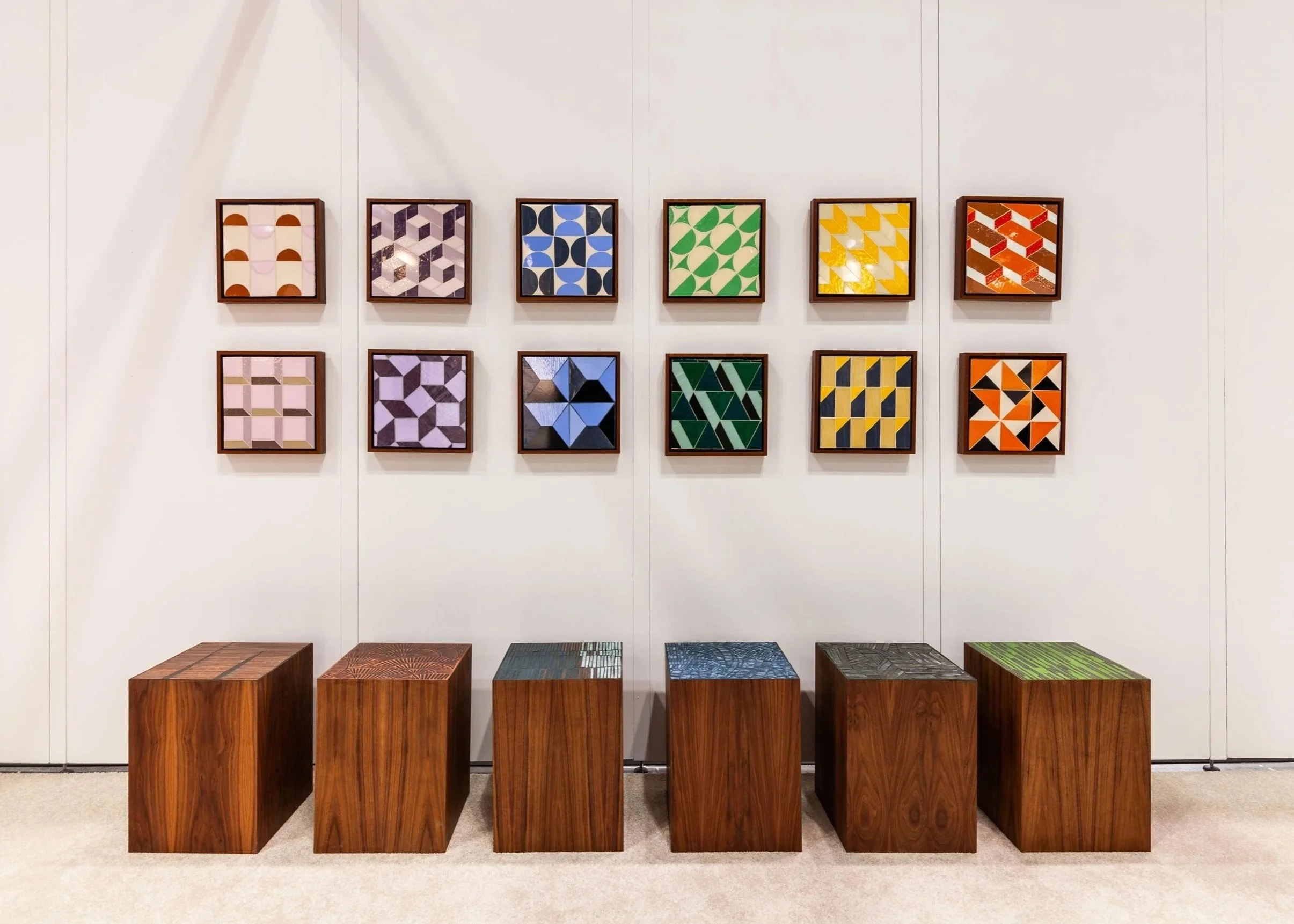 Art gallery wall displaying twelve colorful geometric pattern prints in frames, six at the top and six at the bottom, with six wooden stools in front.
