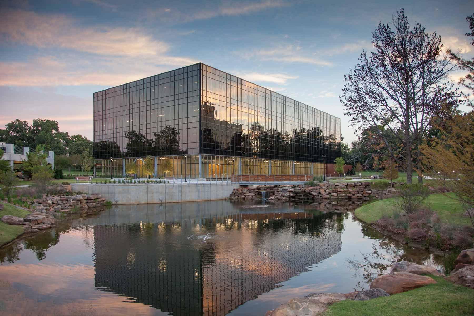 Modern glass office building reflecting the sky and trees, with a pond and landscaped greenery in the foreground.