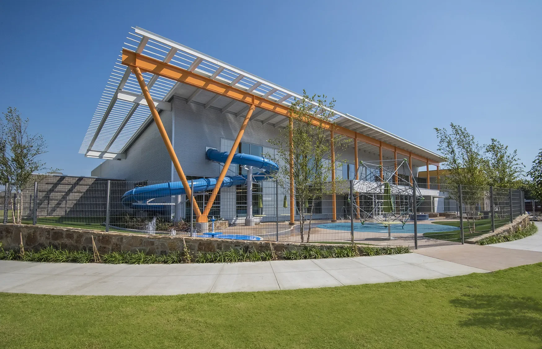 Modern building with a blue water slide and a playground area enclosed by a fence, surrounded by a grassy lawn and trees under a clear sky.