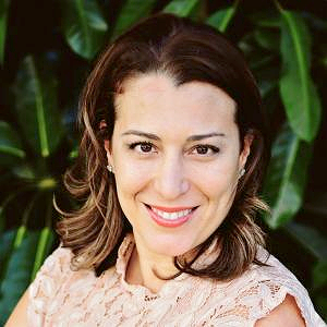 A woman with shoulder-length brown hair smiling in front of green plants.
