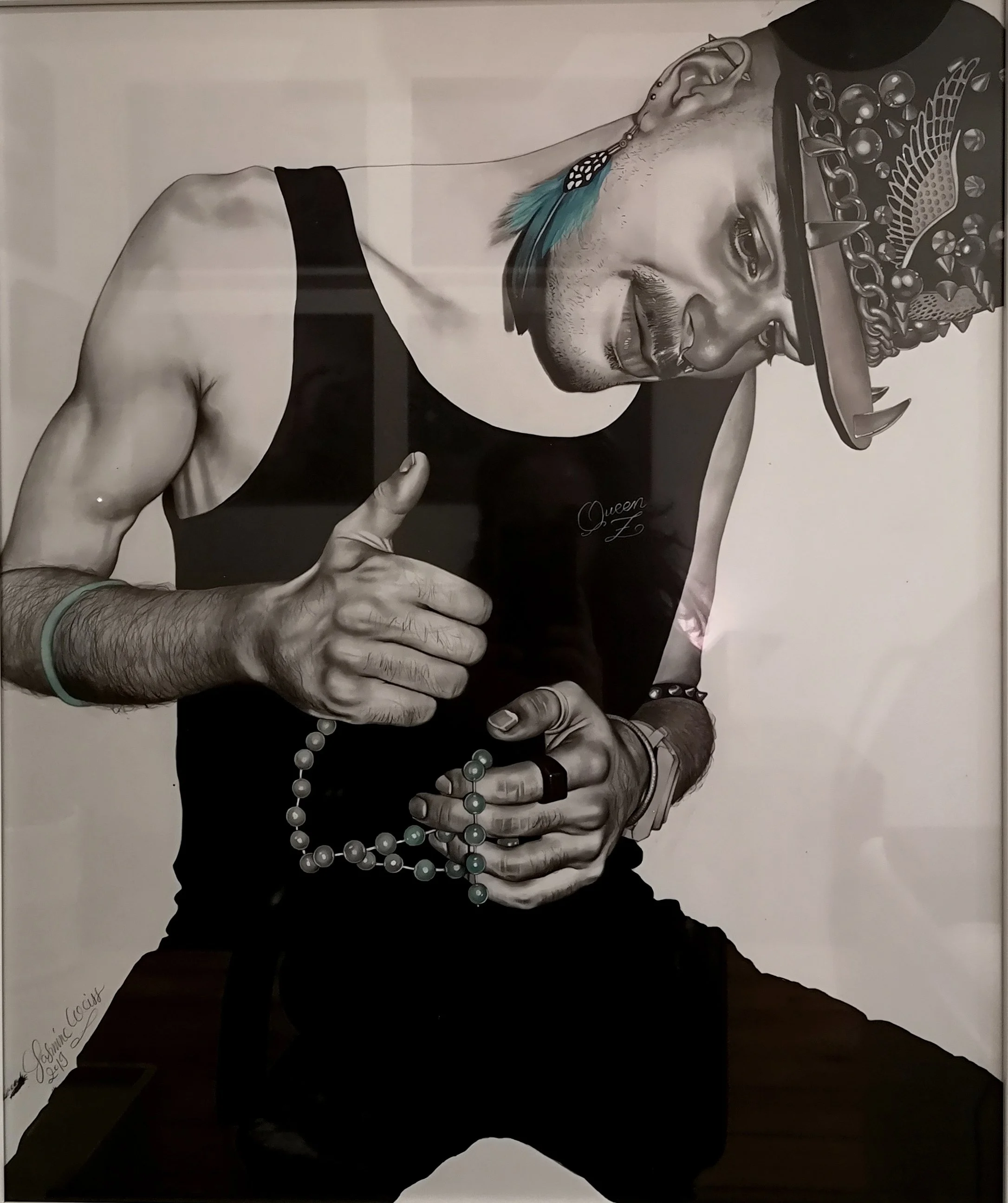 Black and white portrait of a man wearing a detailed headdress, a necklace with beads, and jewelry. He is dressed in a sleeveless shirt with the word 'Queen' visible and is holding a string of beads. The man is smiling and giving a thumbs-up with his