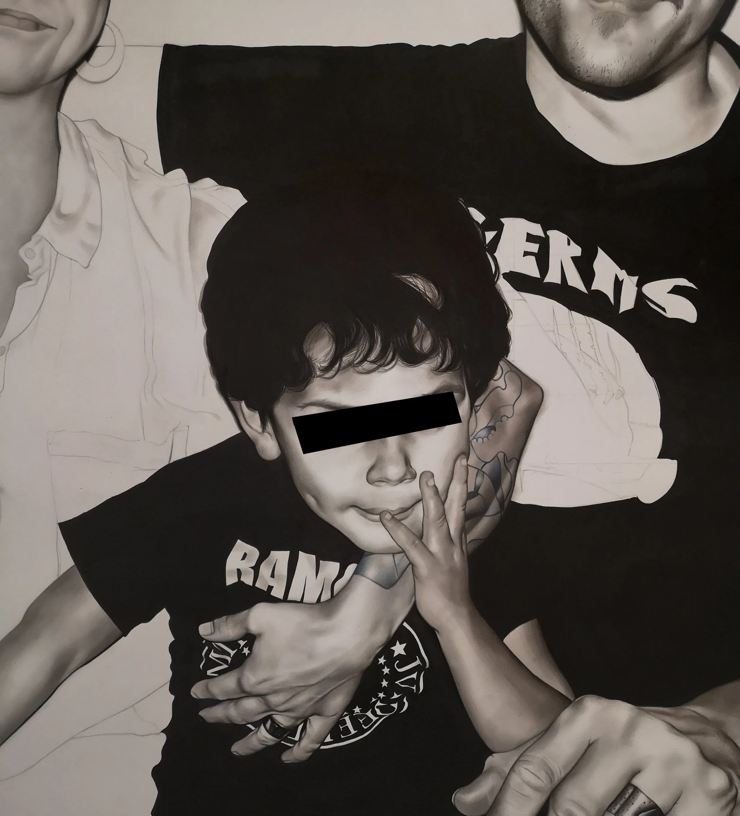 A black and white portrait of a young boy with a relaxed expression, holding a finger to his lips, surrounded by two adults, with their lower faces and arms visible. The child is wearing a Rammstein T-shirt.
