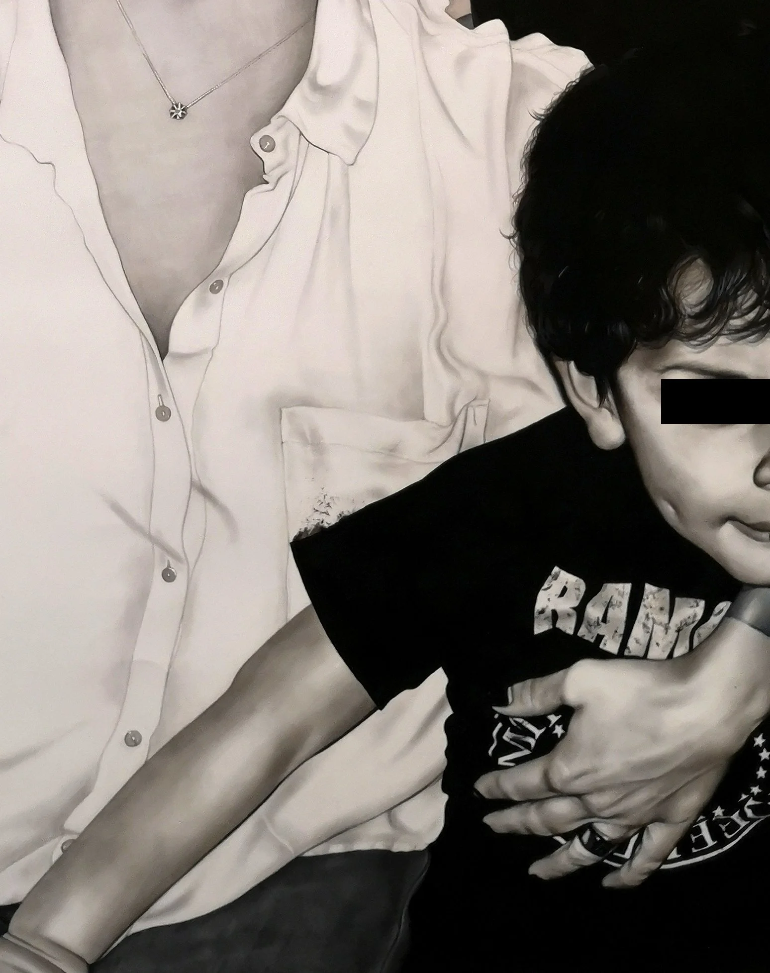 A person with black hair and glasses wearing a black T-shirt with white text, held by another person wearing a white shirt with a necklace, with part of the person's arm visible.