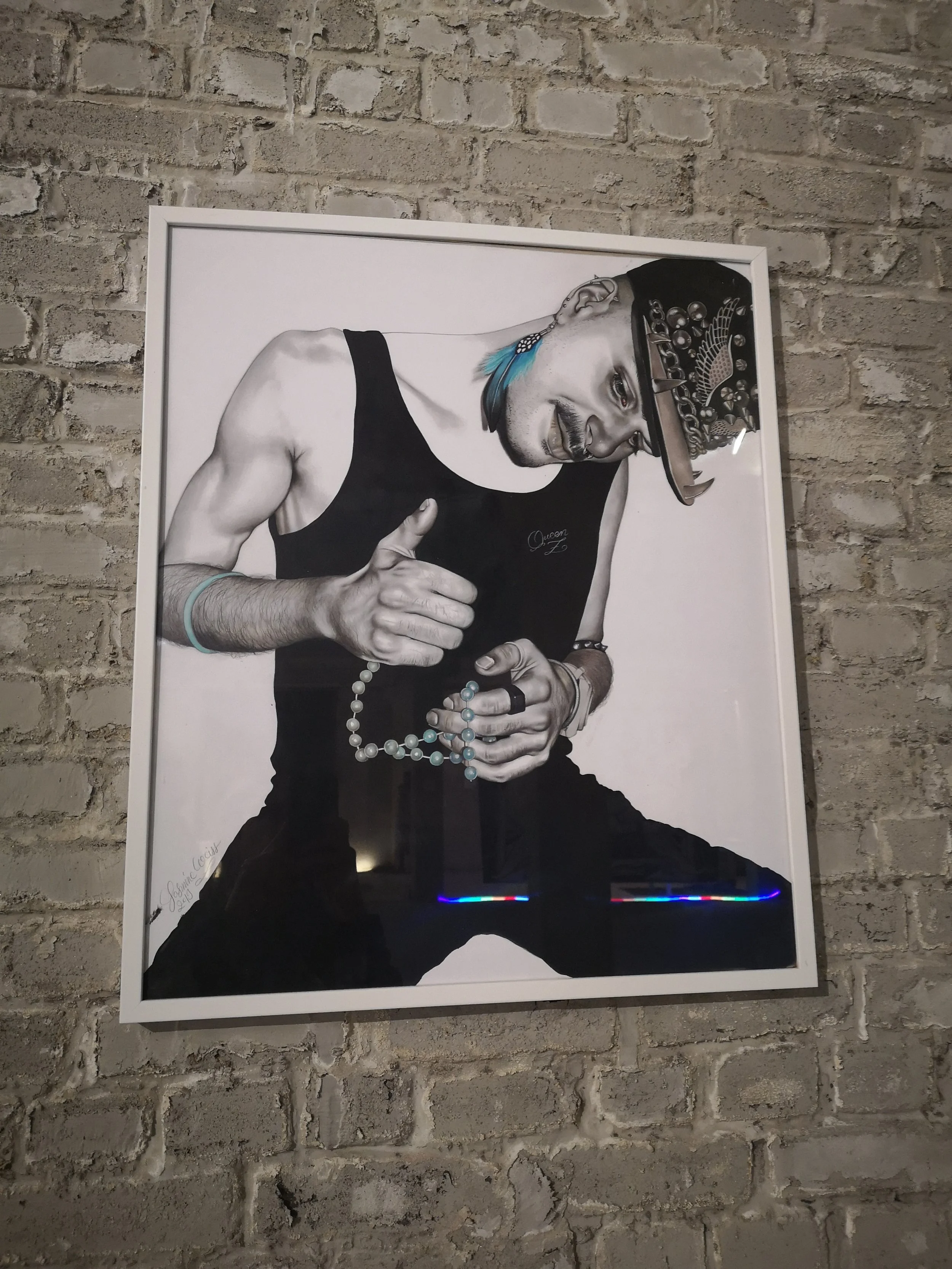 A black and white portrait of a man with a muscular arm, wearing a sleeveless shirt and various jewelry, including a beaded necklace and a bracelet. He is looking to the side and wearing a decorated hat. The portrait is framed and displayed on a bric