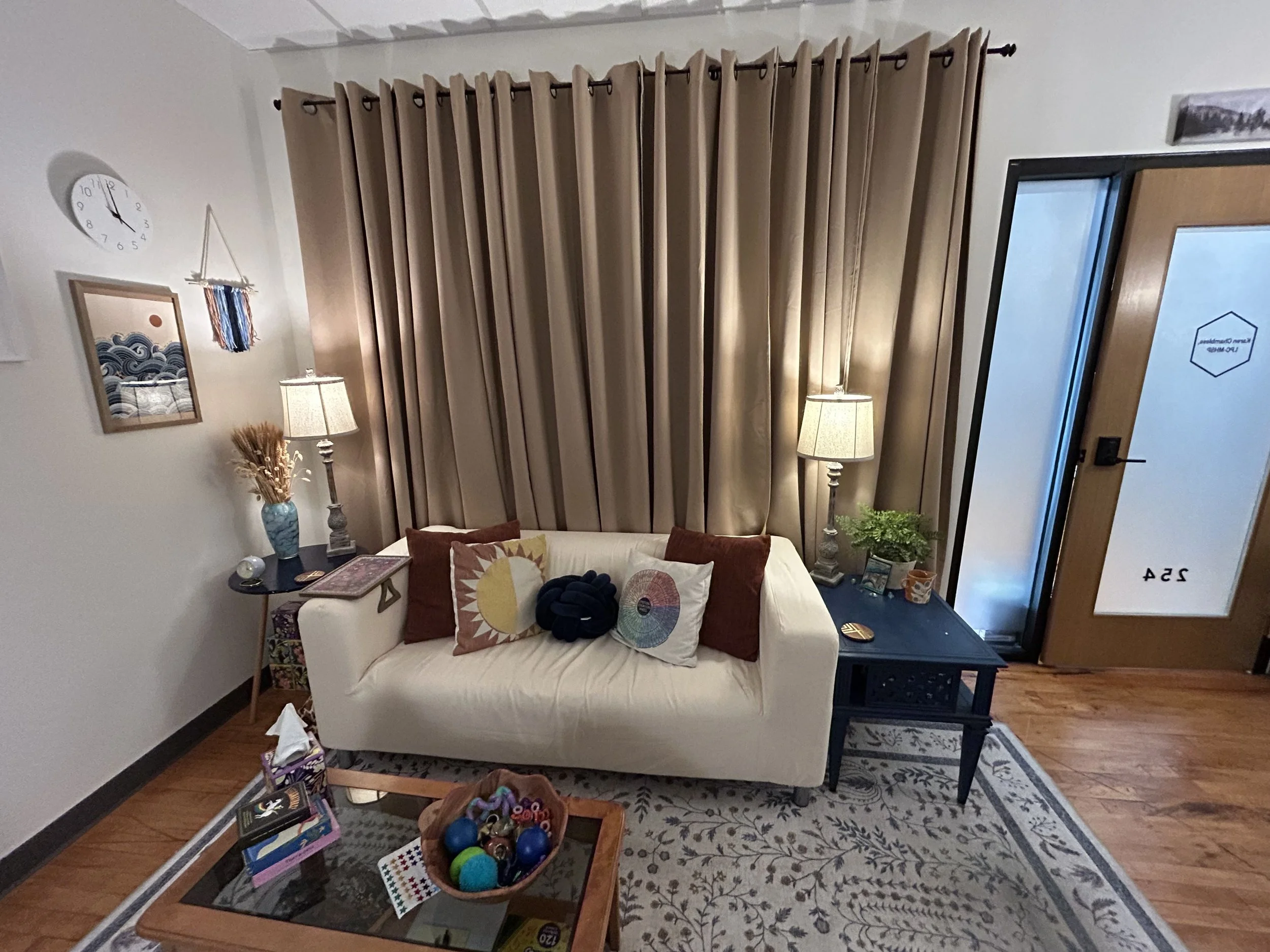 The couch in Karen's therapy office, with lamps on either side, curtains behind the couch, and a coffee table with tissues and a variety of fidget toys.