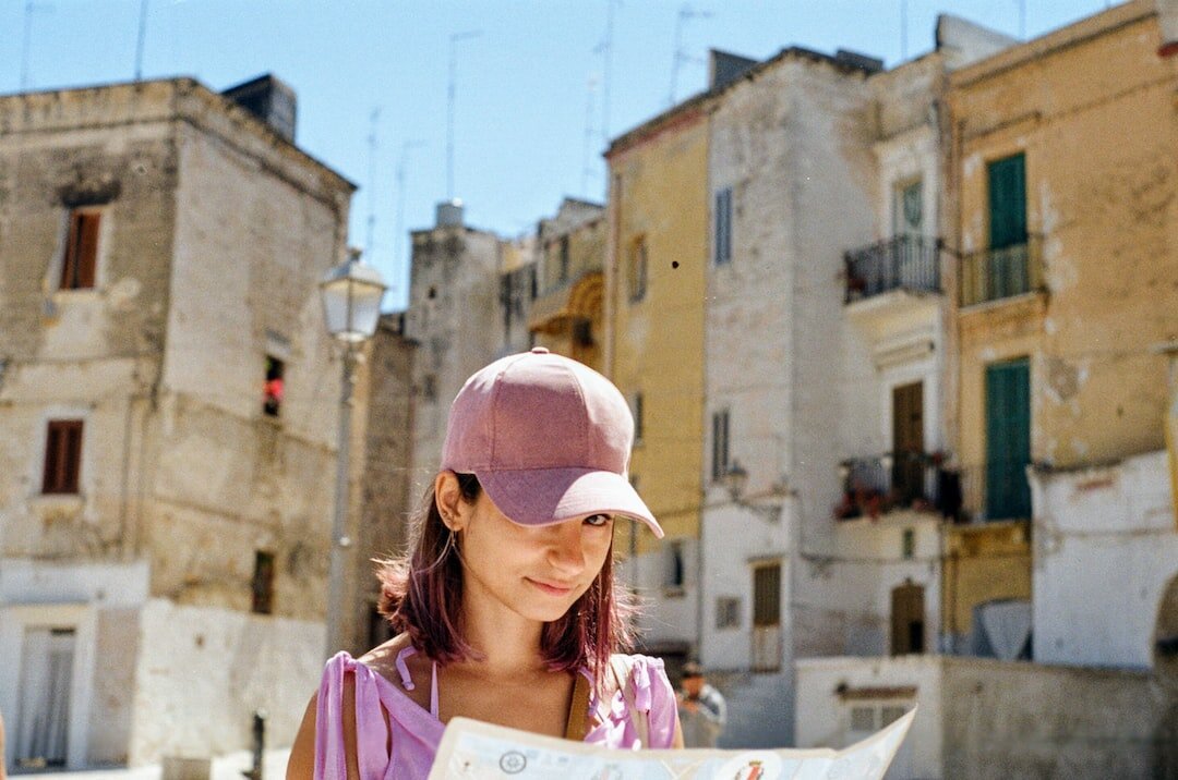 A woman wearing a baseball cap and holding a map looks up at the camera. Behind her are old buildings and blue sky.
